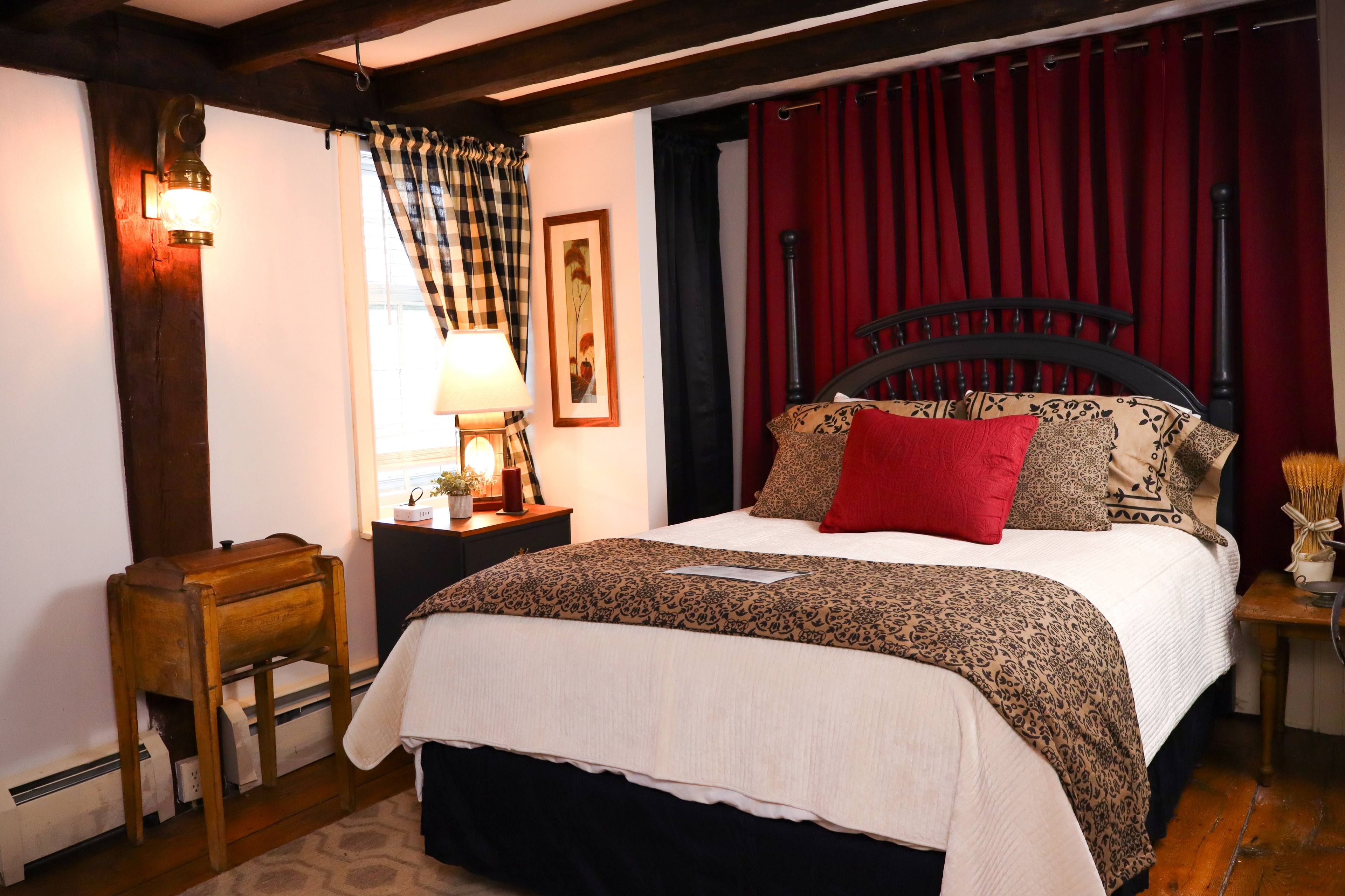 A warm cozy bedroom with dark wood exposed beams, a white quilt with a primitive style blanket, black nightstand and burgundy and plaid drapes