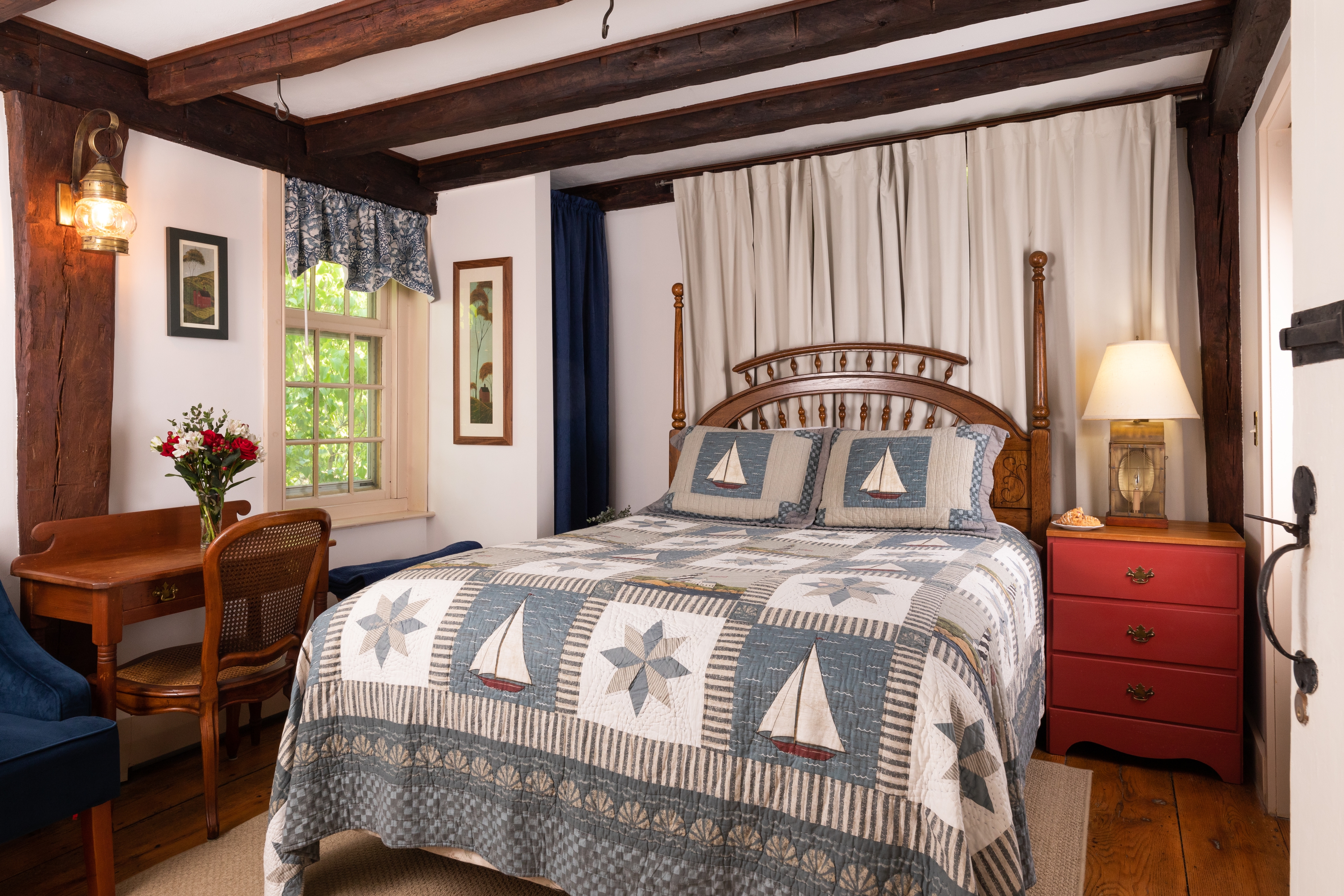 A quaint, nautical-themed bedroom with dark wood beams, a sailboat quilt, a writing desk, and a bright red nightstand.