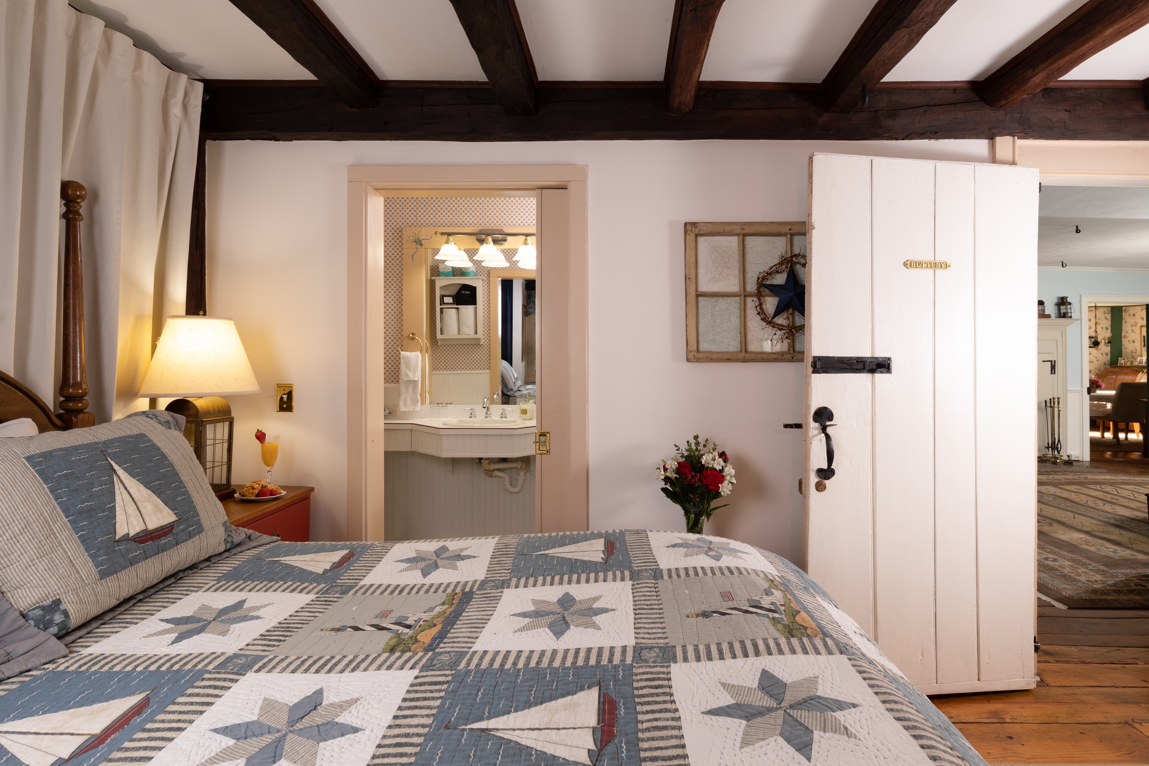 A charming, nautical bedroom with a sailboat quilt, exposed dark wood beams, and an open doorway leading to a brightly lit bathroom.