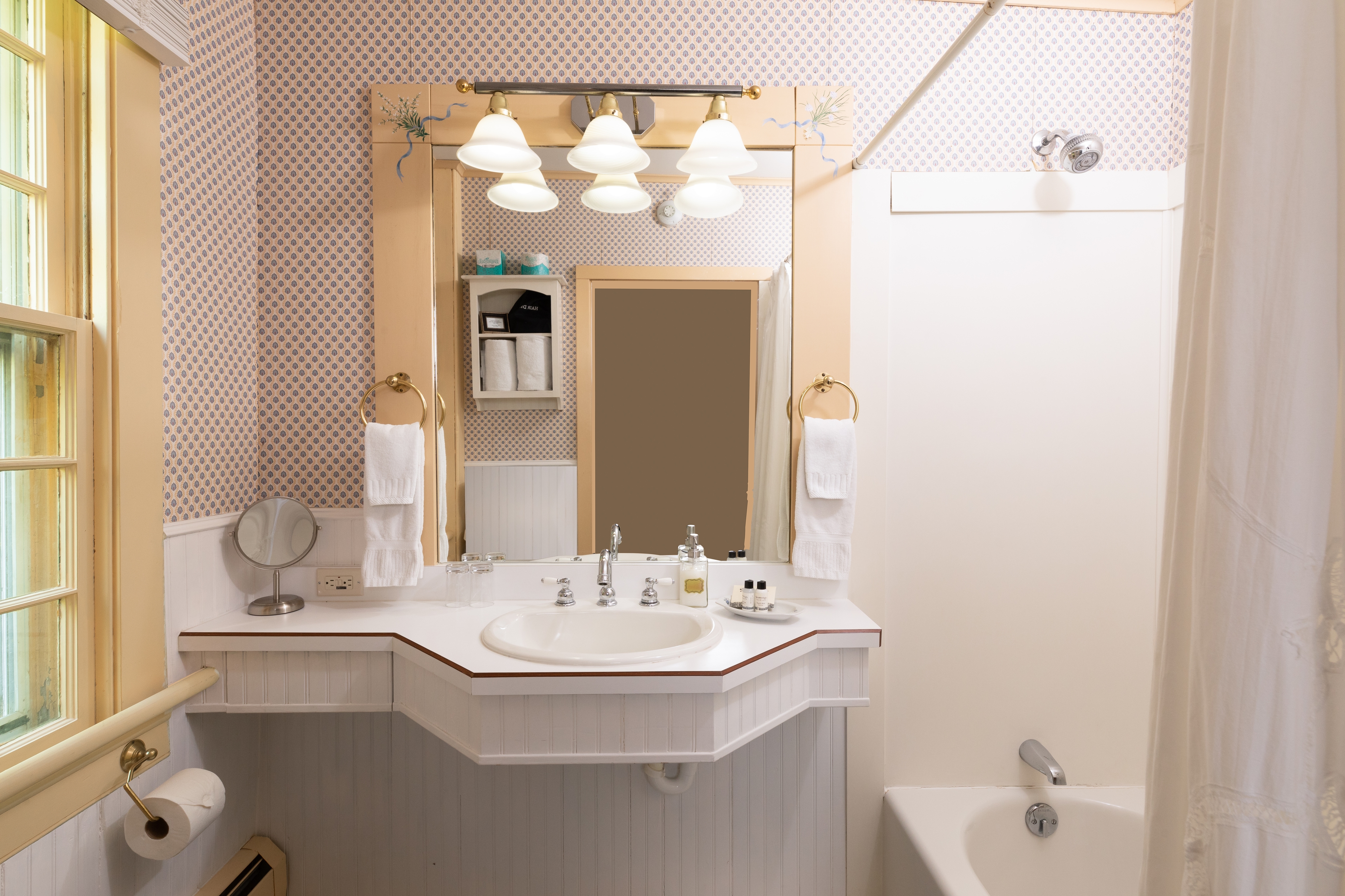 A quaint, brightly lit bathroom with a unique angled vanity, a large mirror with decorative lighting, and a standard shower/tub combo.