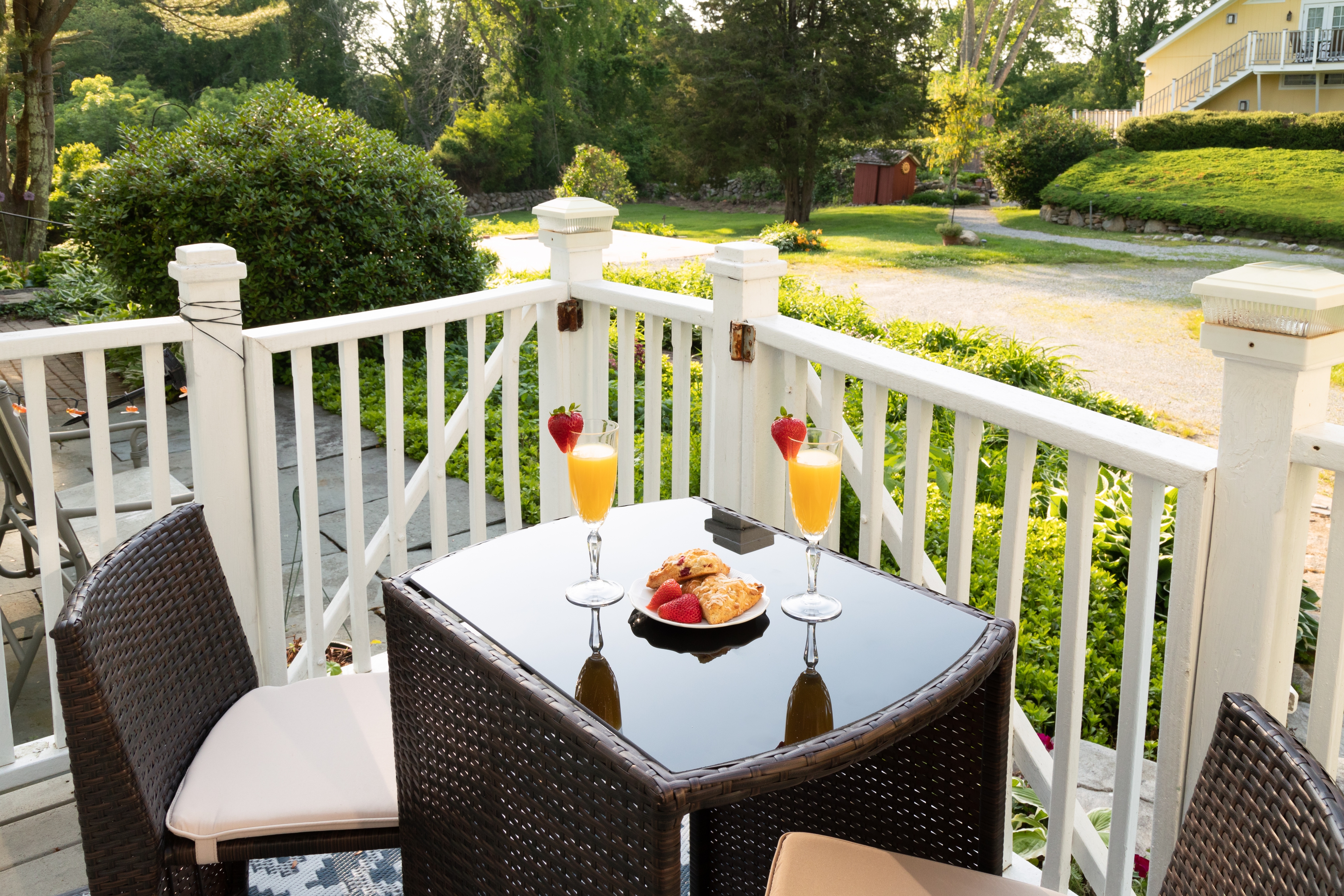 A bright, inviting outdoor balcony with a wicker table set for two, featuring mimosas and pastries, overlooking a sunny garden area.