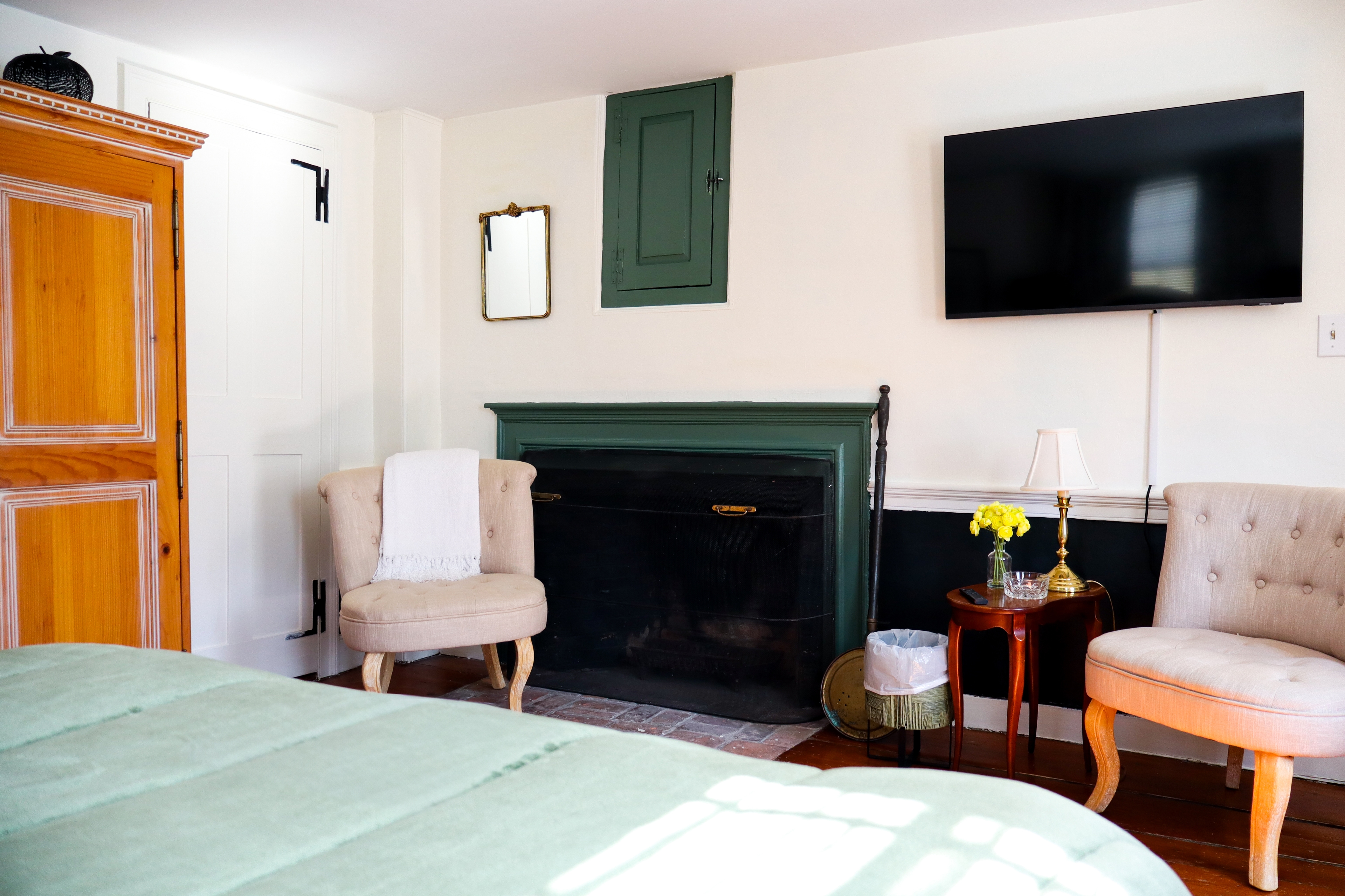 A bright, cozy bedroom with a large bed, light wood armoire, and an open doorway revealing a pedestal sink and mirror in the adjacent bathroom.