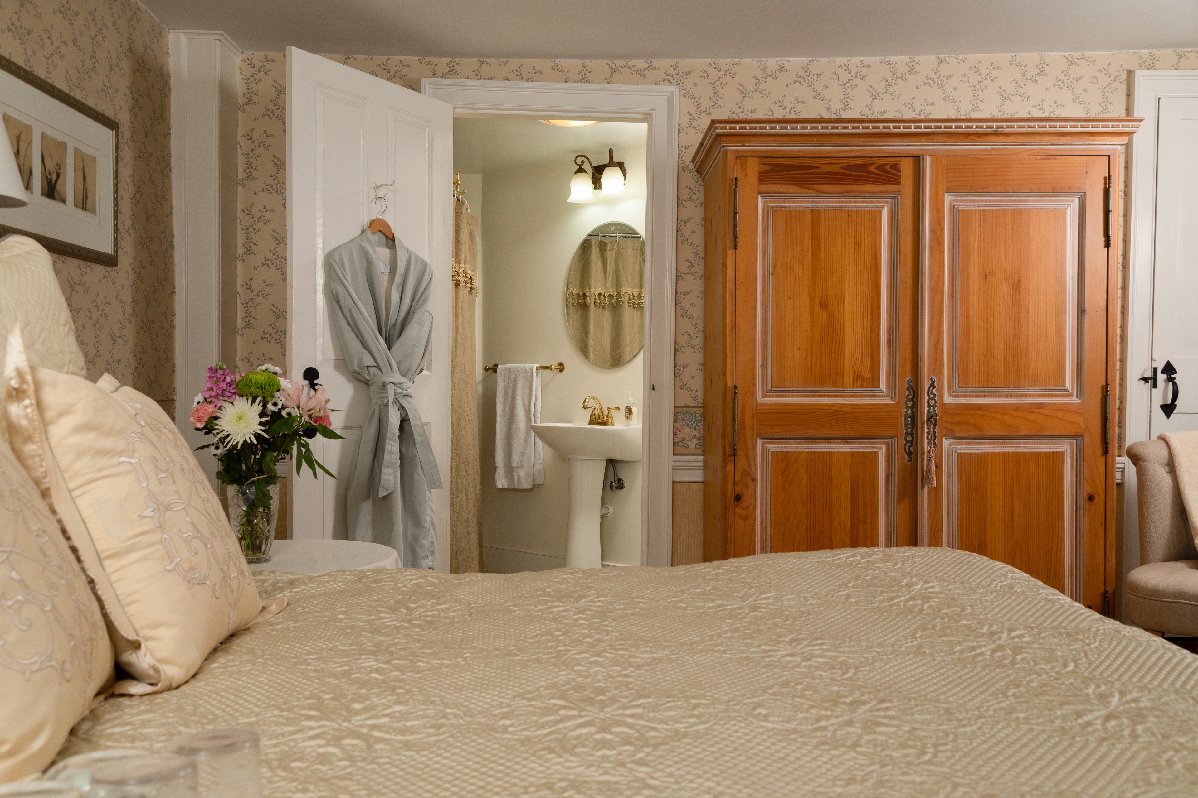 A bright, cozy bedroom with a large bed, light wood armoire, and an open doorway revealing a pedestal sink and mirror in the adjacent bathroom.