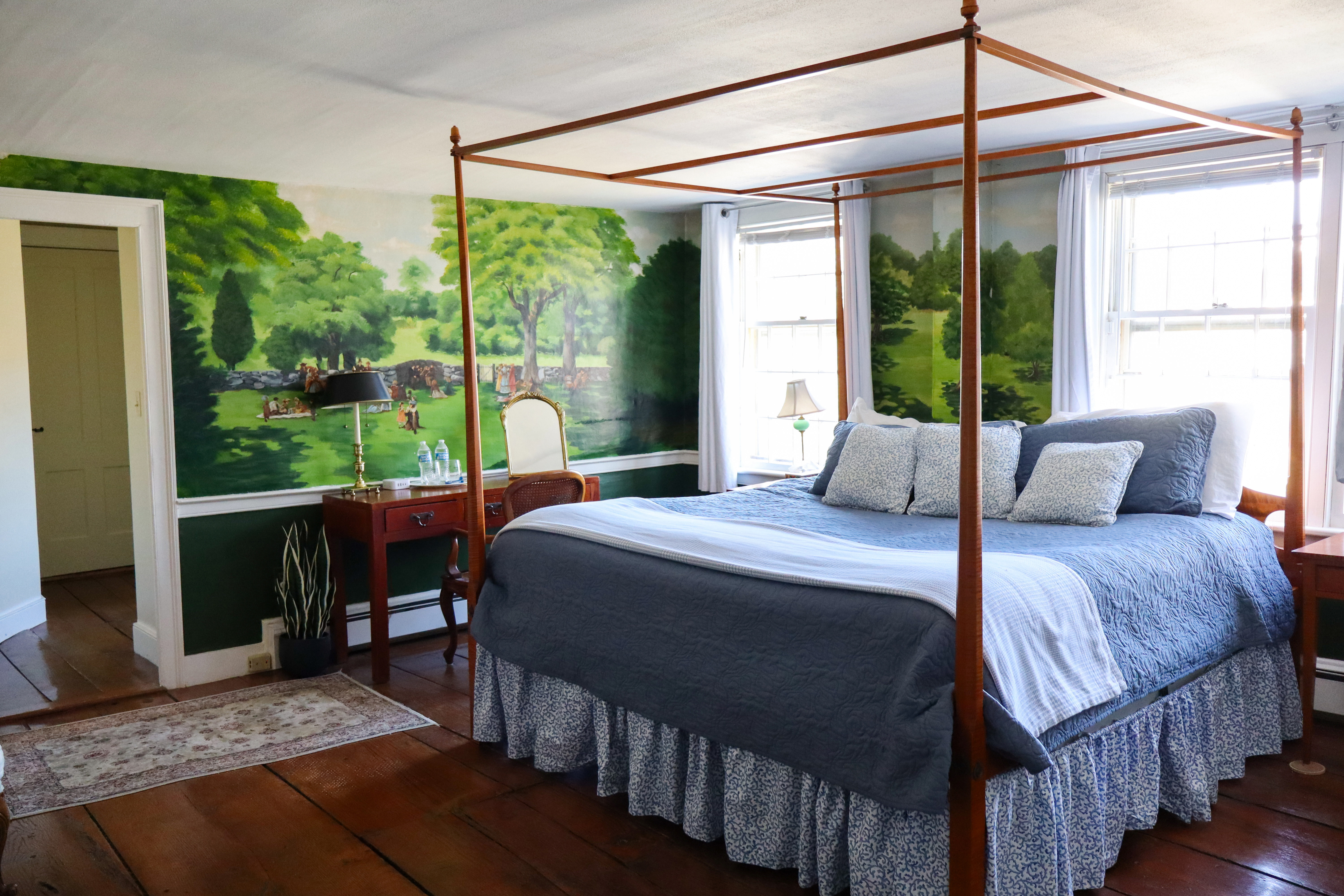 An enchanting, romantic bedroom featuring a hand-painted landscape mural behind a cozy, lit fireplace and a canopy bed with champagne.
