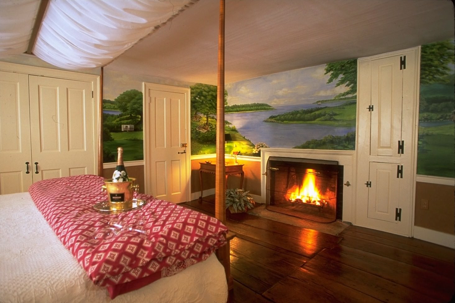An enchanting, romantic bedroom featuring a hand-painted landscape mural behind a cozy, lit fireplace and a canopy bed with champagne.