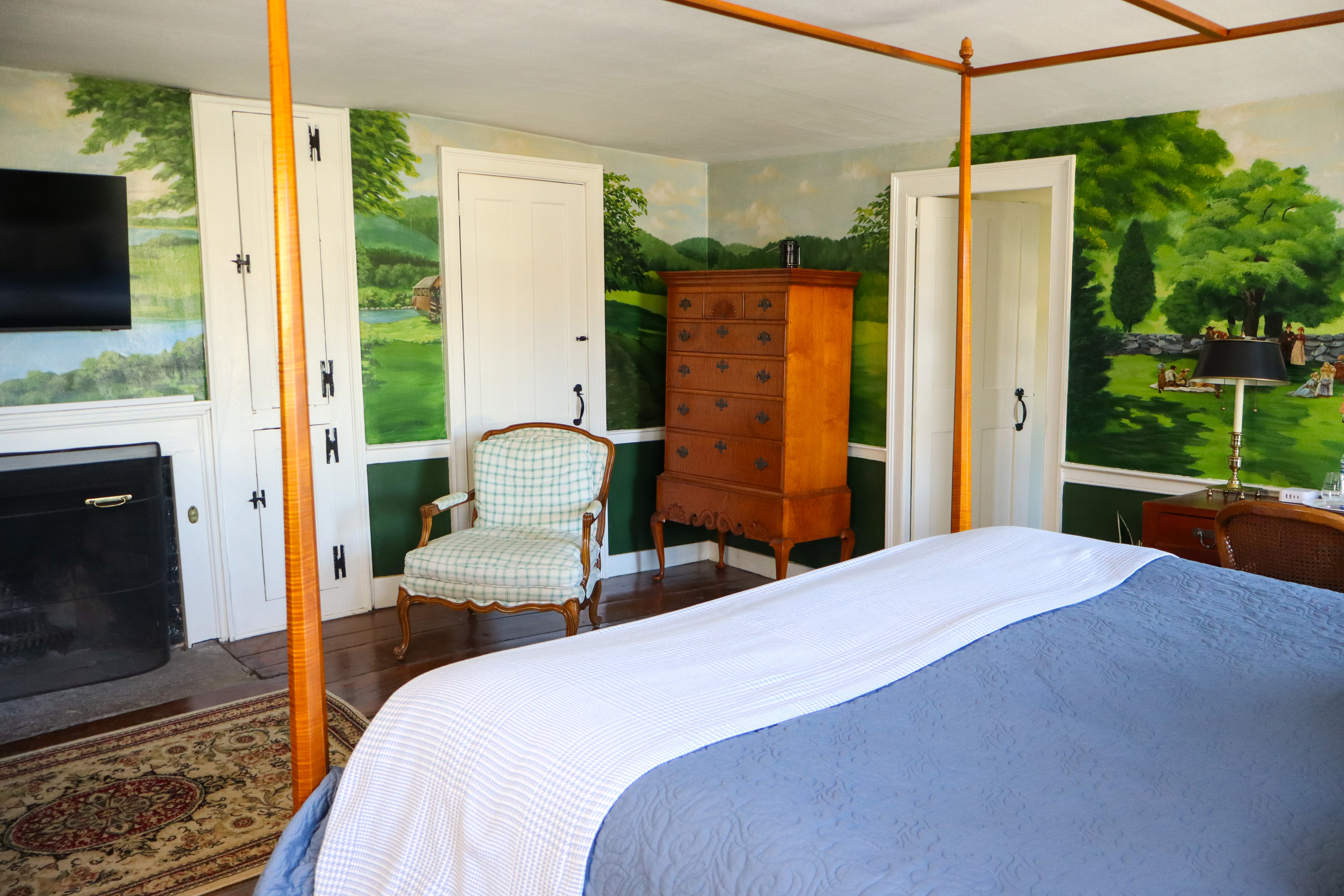 A romantic bedroom scene with a lit fireplace, a plaid ottoman, and a hand-painted landscape mural, creating a cozy and unique ambiance.