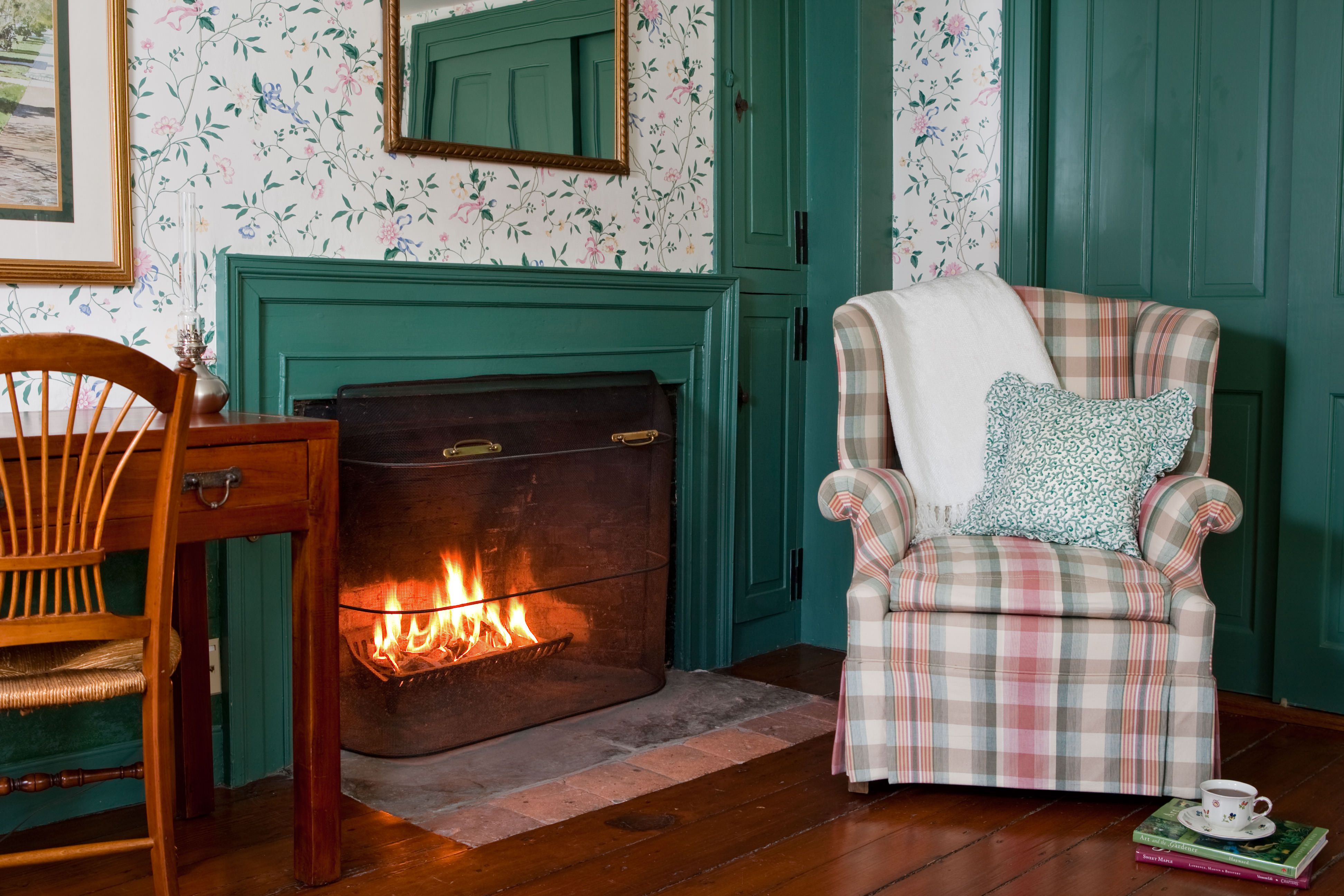 A cozy corner featuring a lit fireplace with a bright green mantel, a plaid armchair, and floral wallpaper, creating a quaint, warm atmosphere.