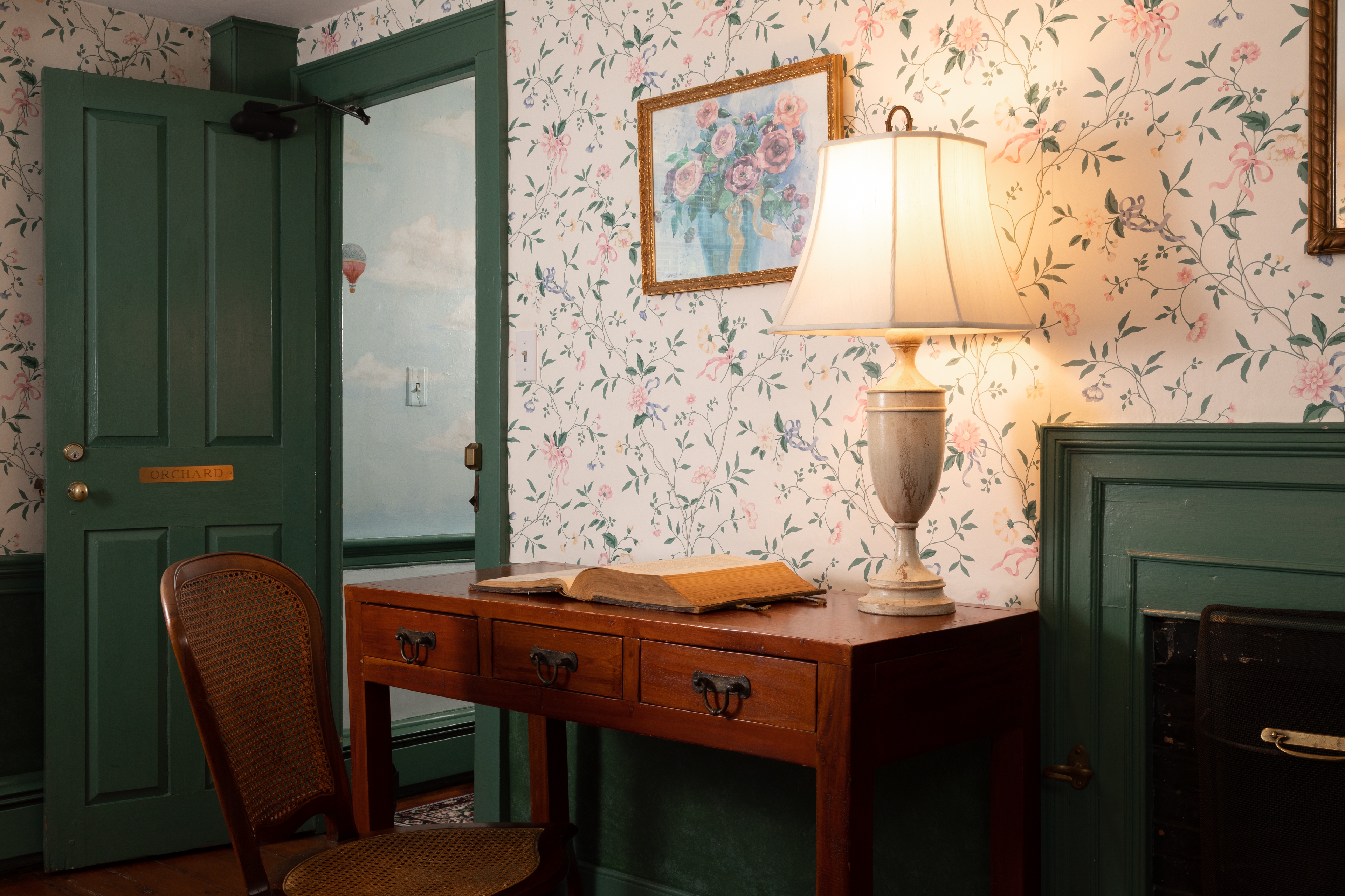 A quaint room featuring delicate floral wallpaper, bright green trim, a small wood writing desk with a lamp, and an open doorway.