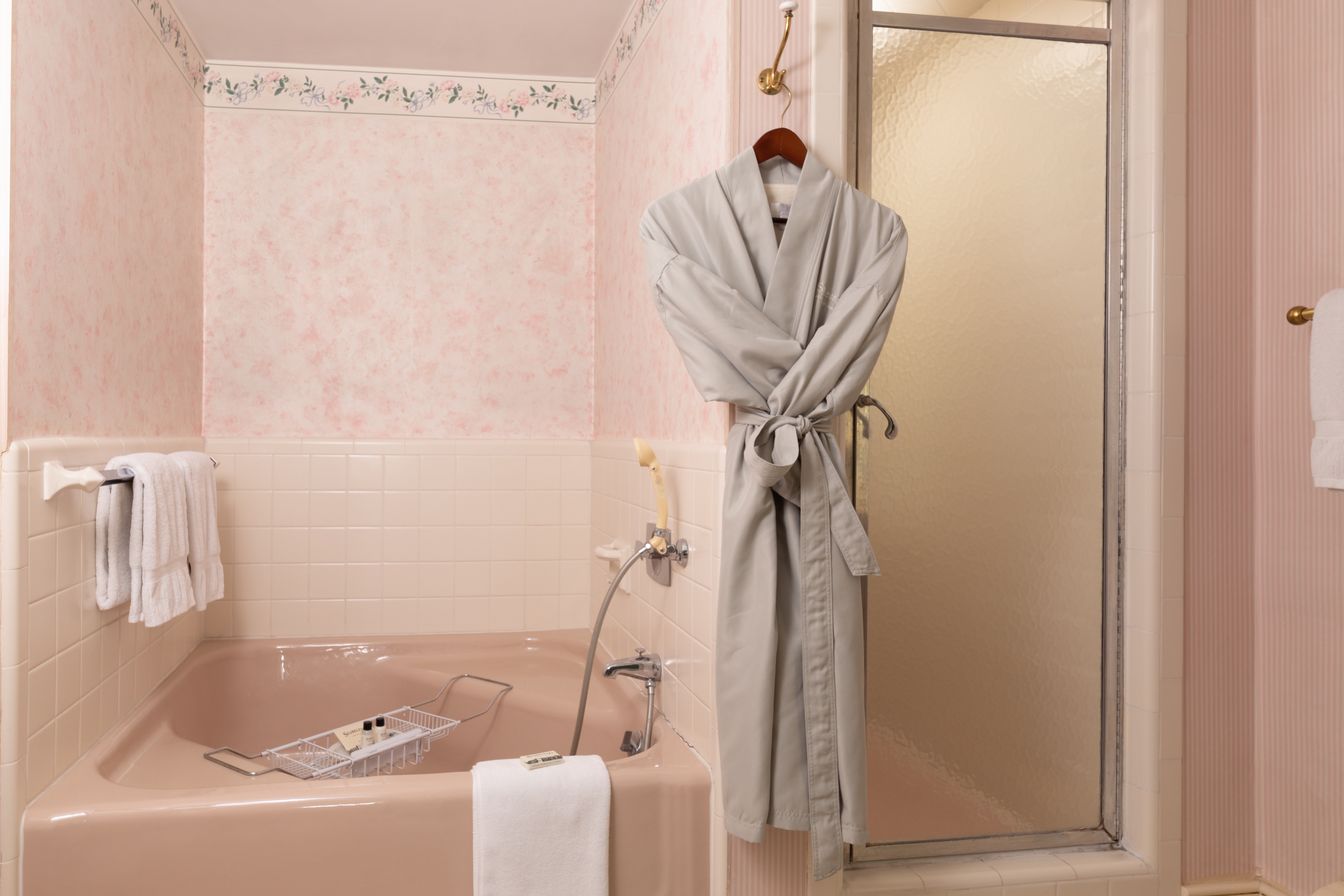 A vintage-style pink bathroom featuring a built-in tub, a glass shower stall, and a cozy gray bathrobe hanging on the wall.