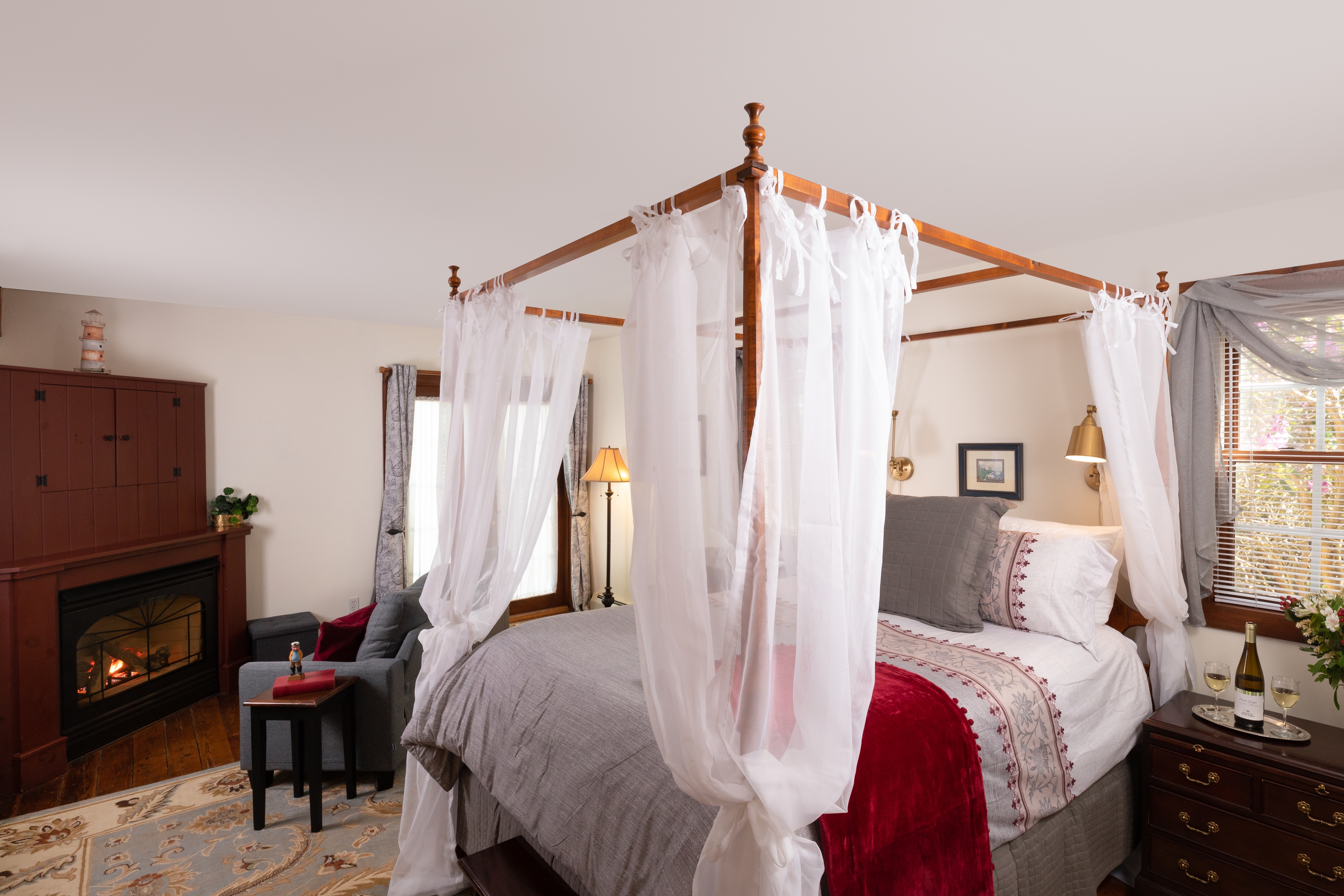 A luxurious bedroom featuring a wooden four-poster bed with sheer white drapes, a cozy fireplace, and a welcoming wine setup.