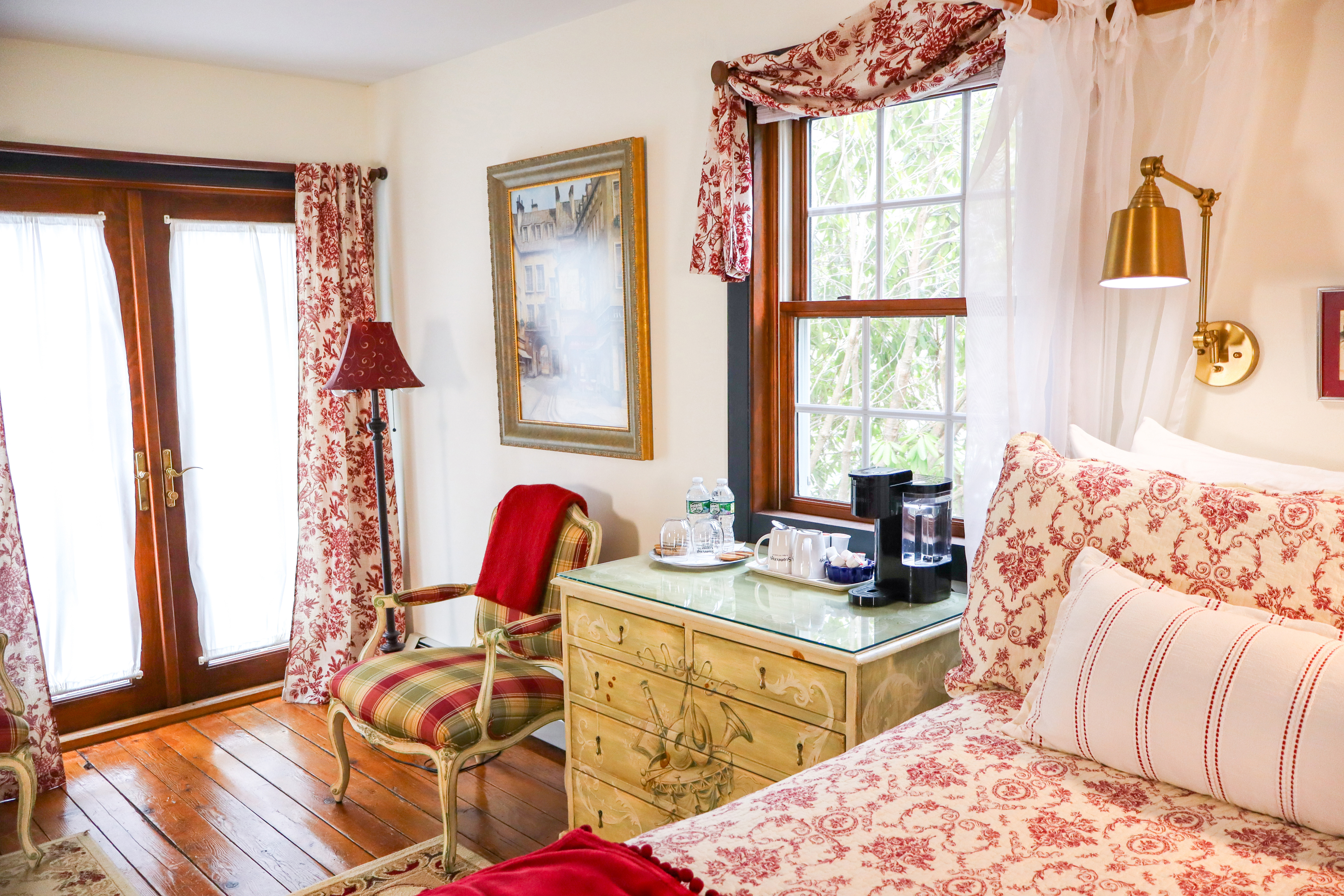 A cozy sitting area with a lit fireplace set in a dark red cabinet, a gray sofa with crimson pillows, and French doors to the right.