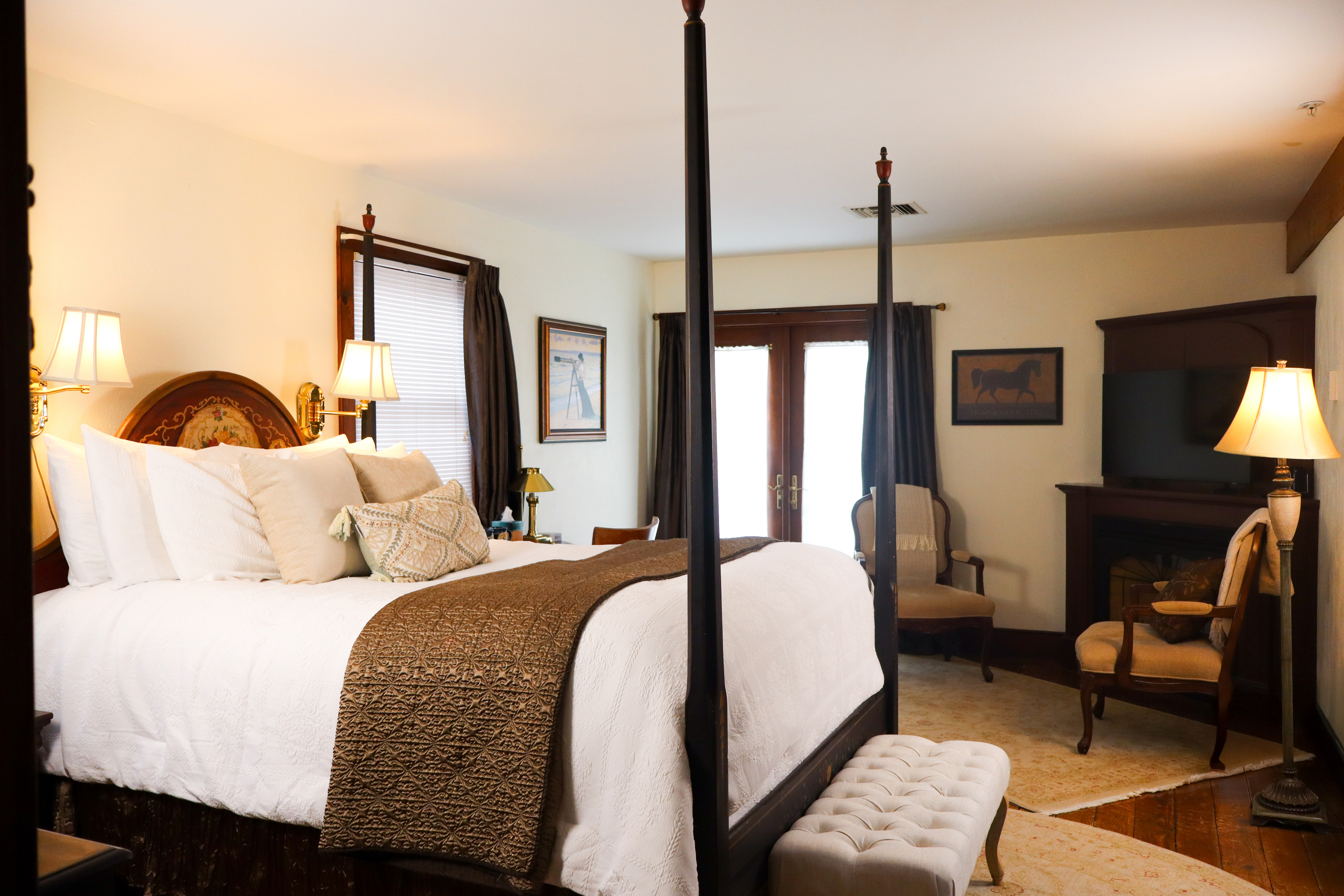 A cozy, elegant bedroom with a dark wood four-poster bed, a fireplace, comfortable armchairs, and floral curtains.