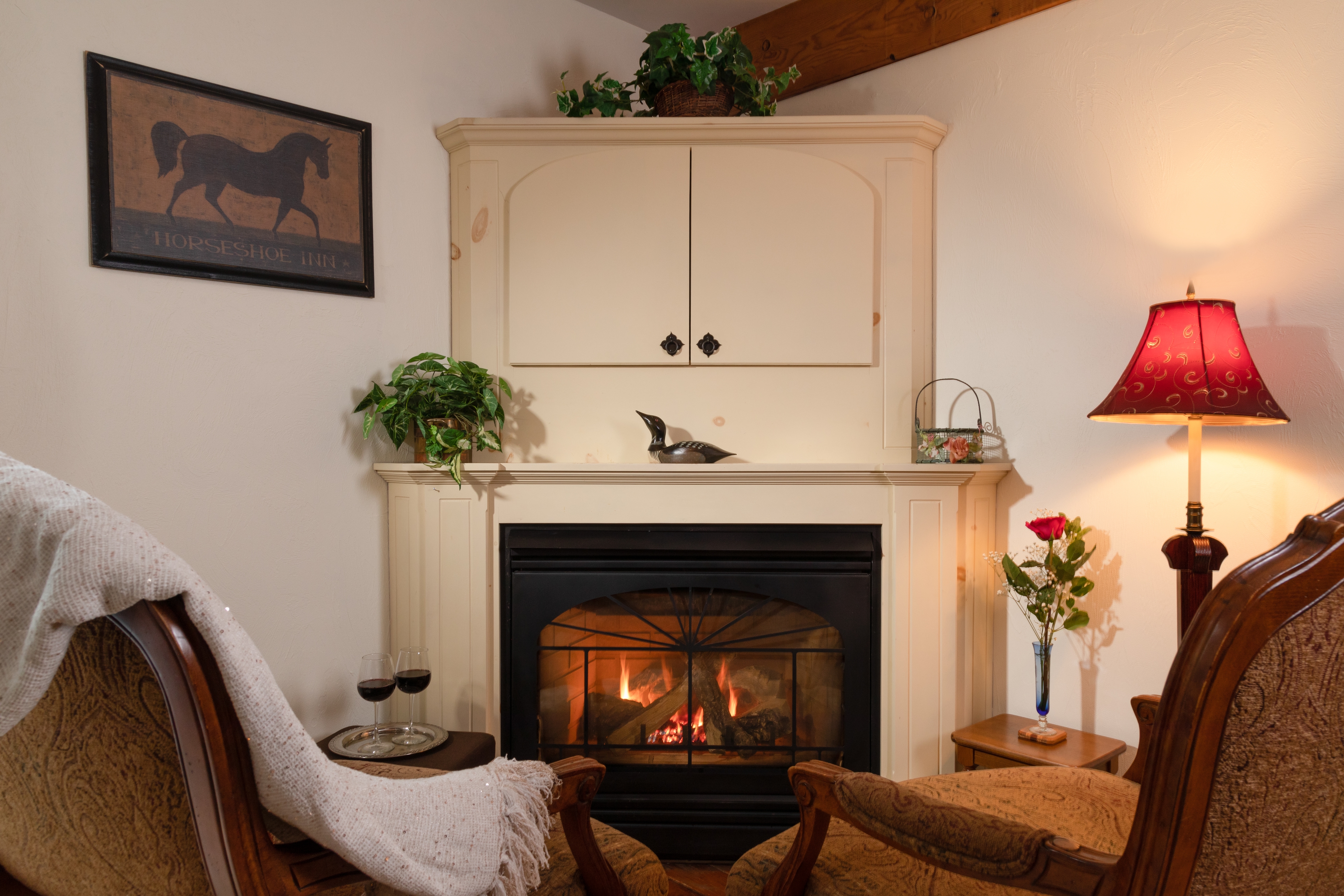 A charming sitting area featuring a lit fireplace, a built-in cabinet mantelpiece, two cozy armchairs, and a tray with wine glasses.
