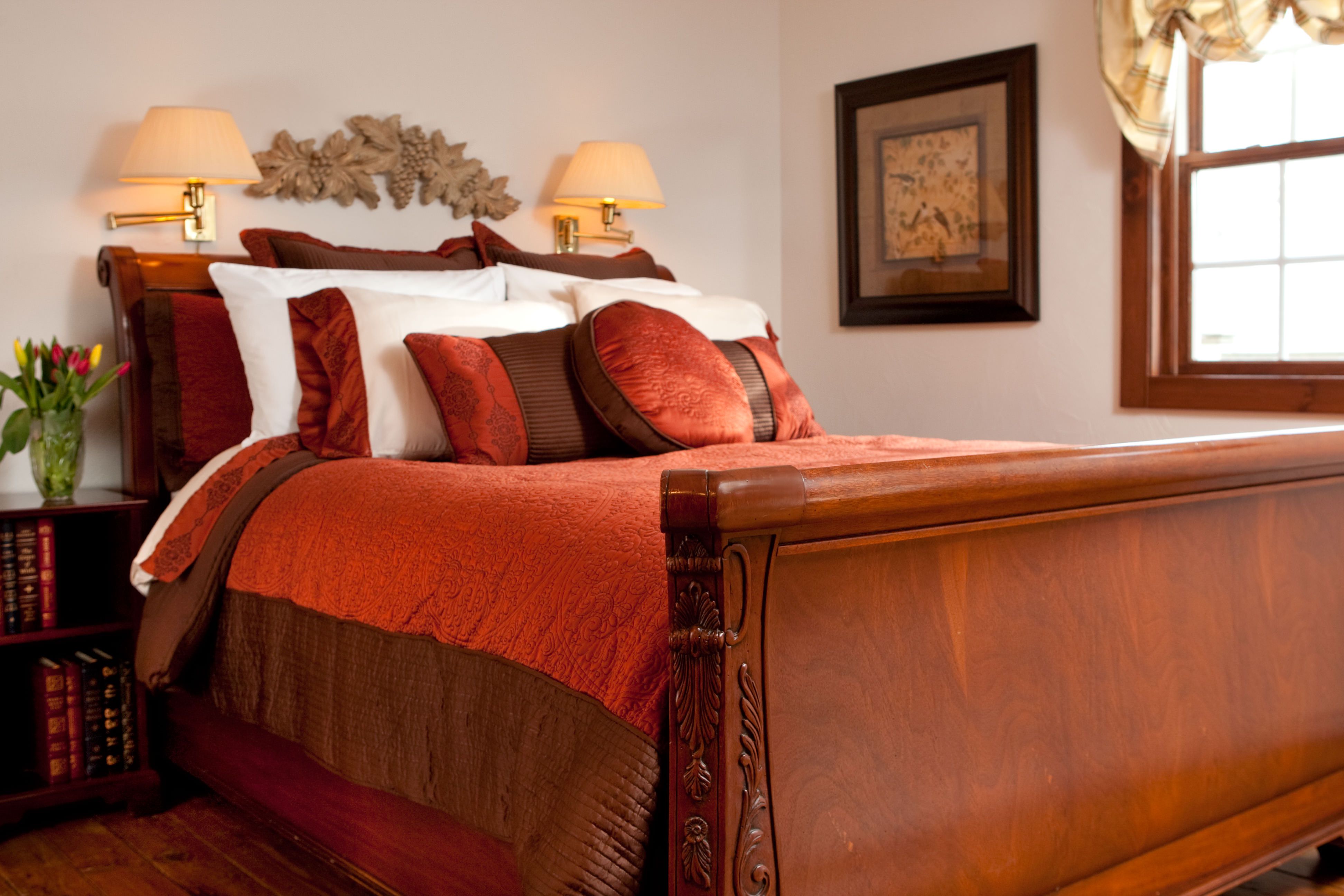 A richly decorated bedroom featuring a warm wooden sleigh bed with burnt orange and brown bedding, illuminated by two bedside lamps.