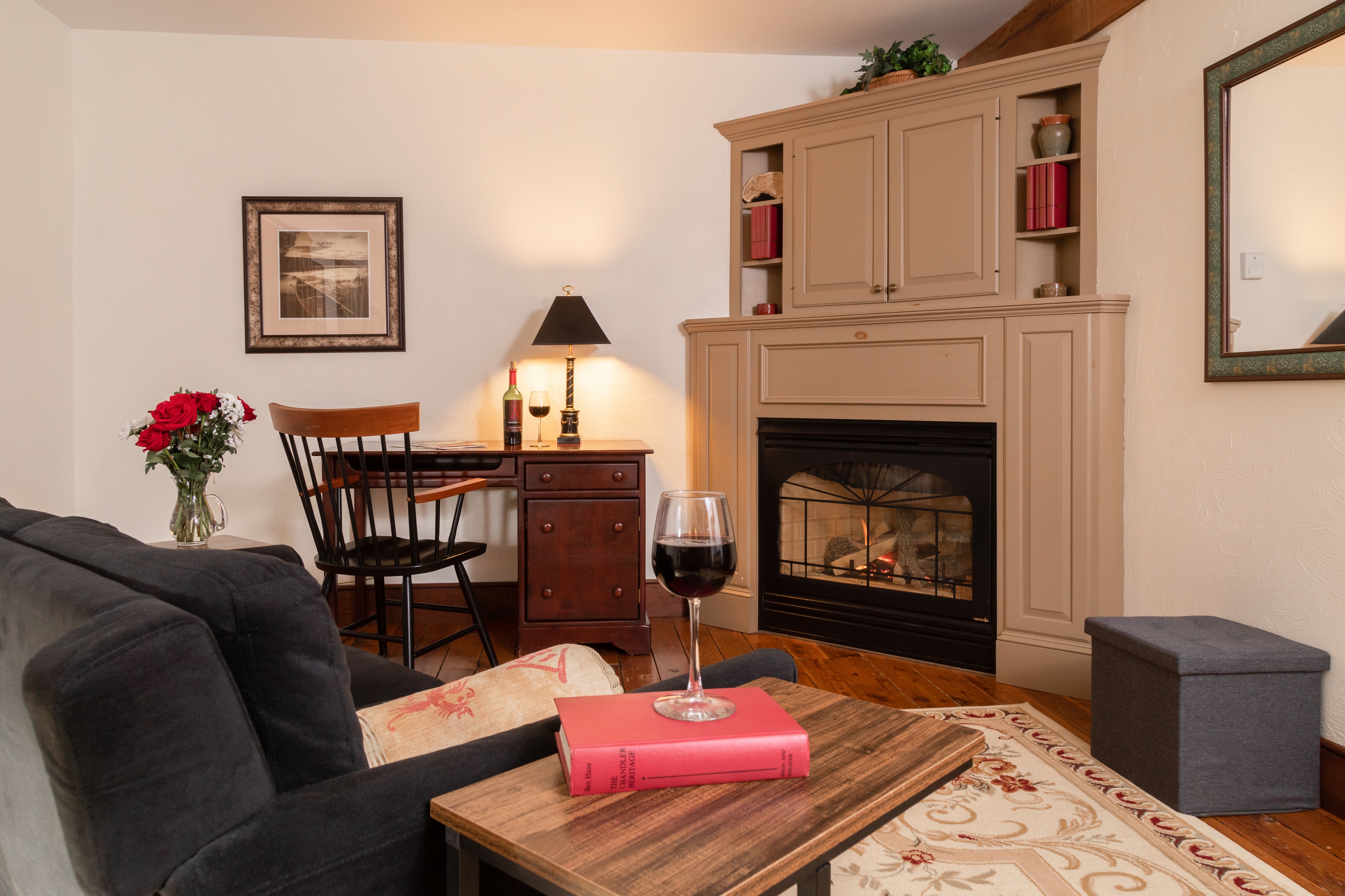 A cozy sitting area with a lit fireplace, a dark blue sofa, a writing desk, and a glass of red wine on a book for a relaxing evening.