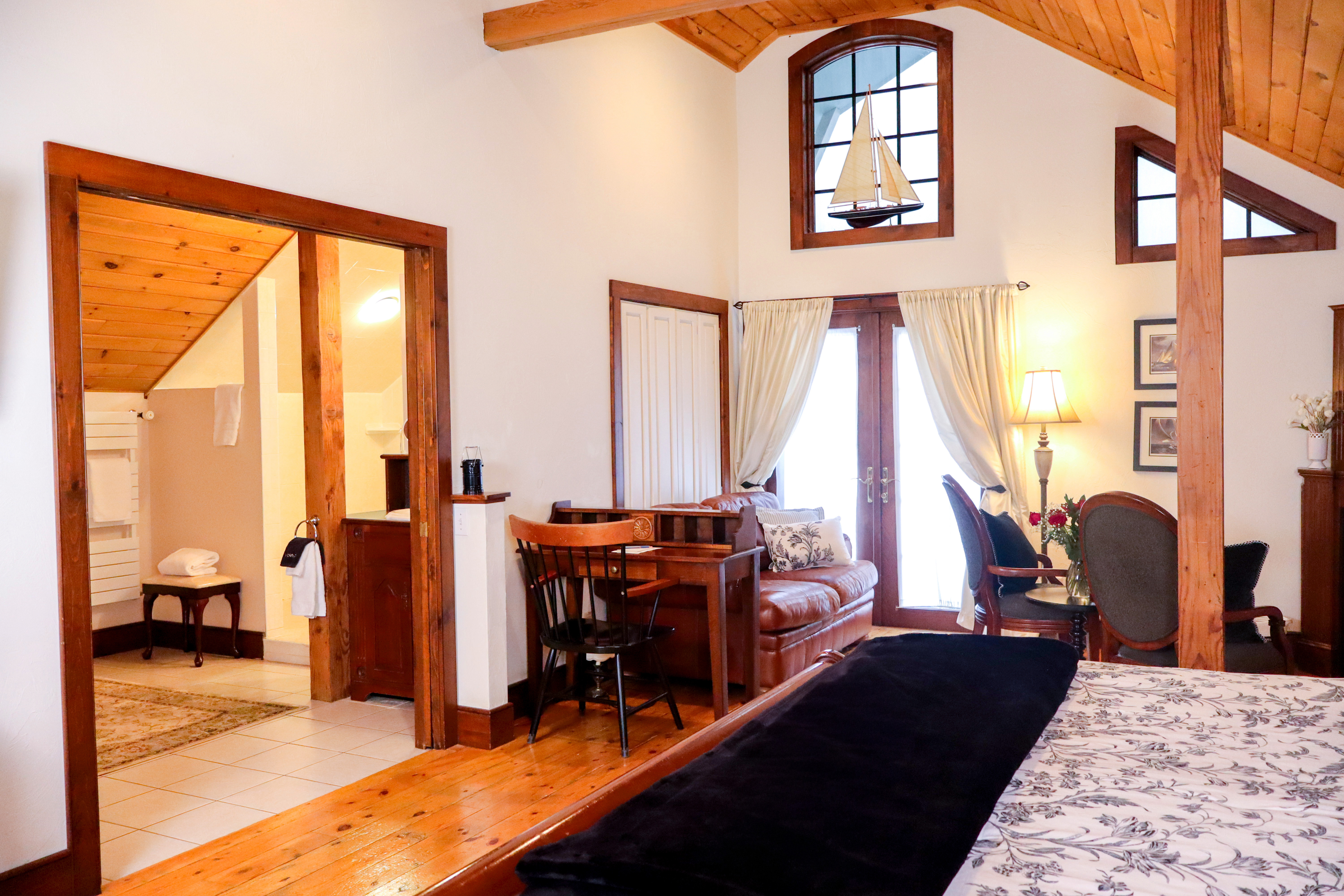 A cozy loft sitting area with a warm wood cathedral ceiling, a lit fireplace in a wood mantel, a brown leather sofa, and French doors.