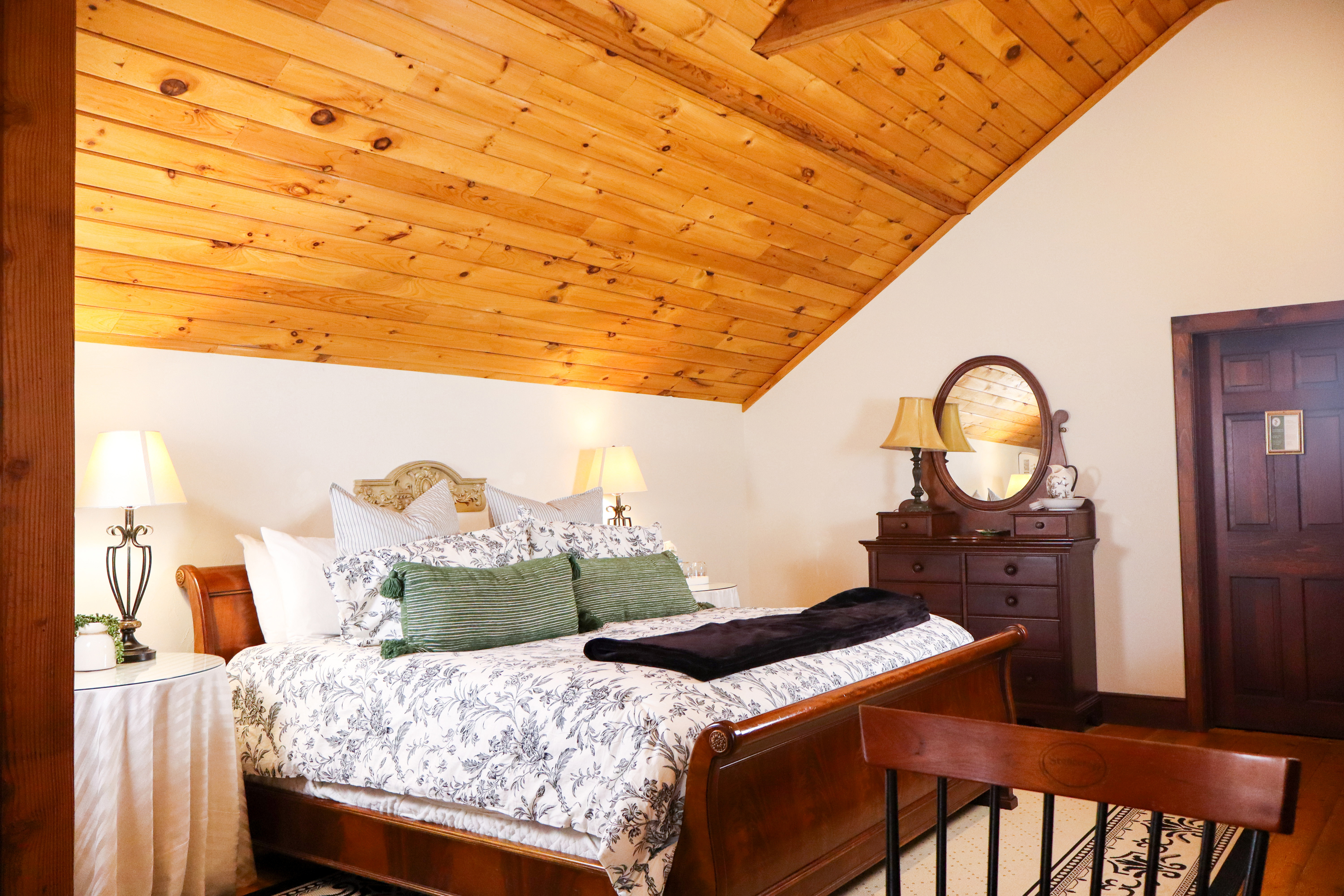 A bedroom with a warm wood cathedral ceiling, a dark wood sleigh bed, and a dresser with an antique mirror.