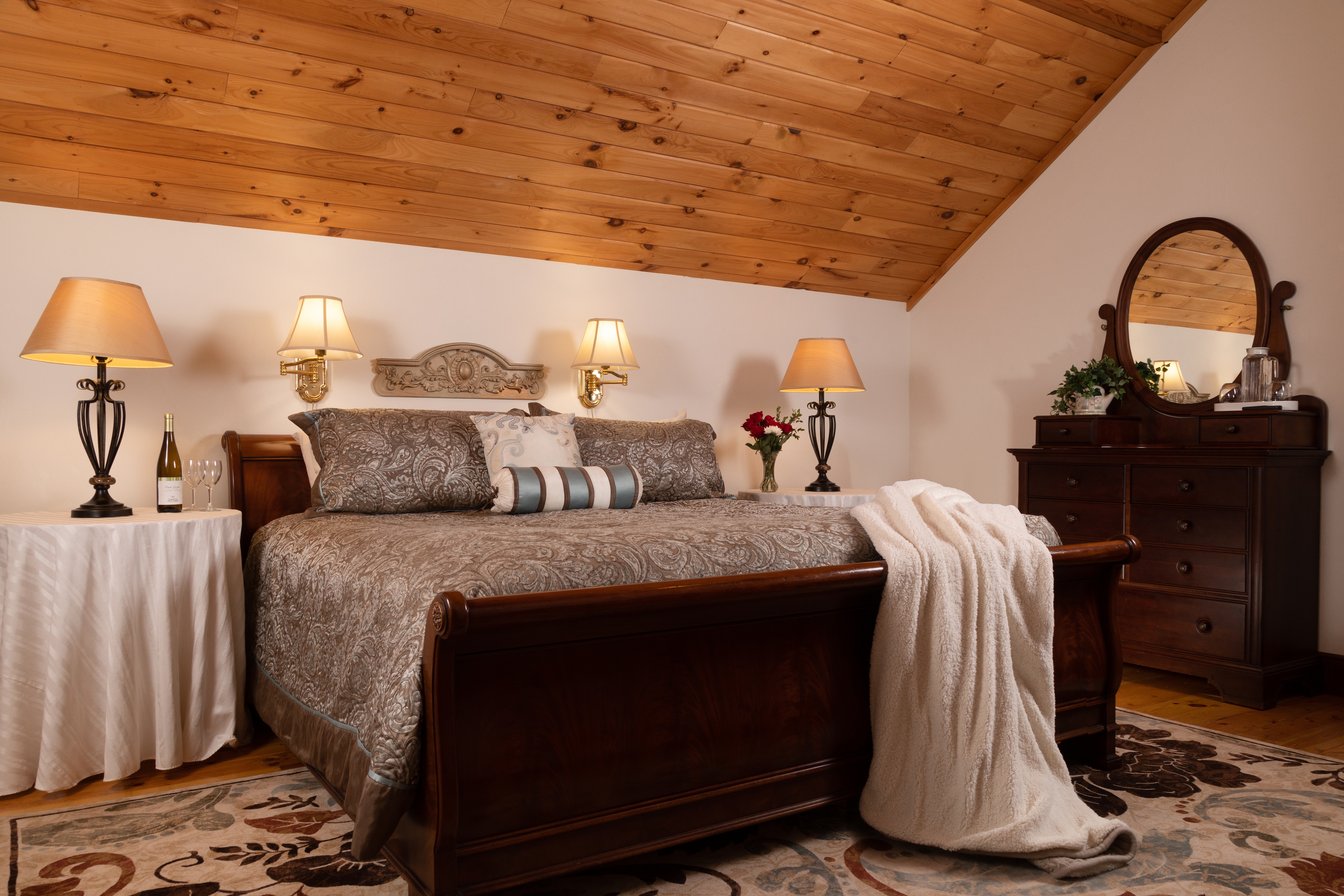 A bedroom with a warm wood cathedral ceiling, a dark wood sleigh bed, and a dresser with an antique mirror.