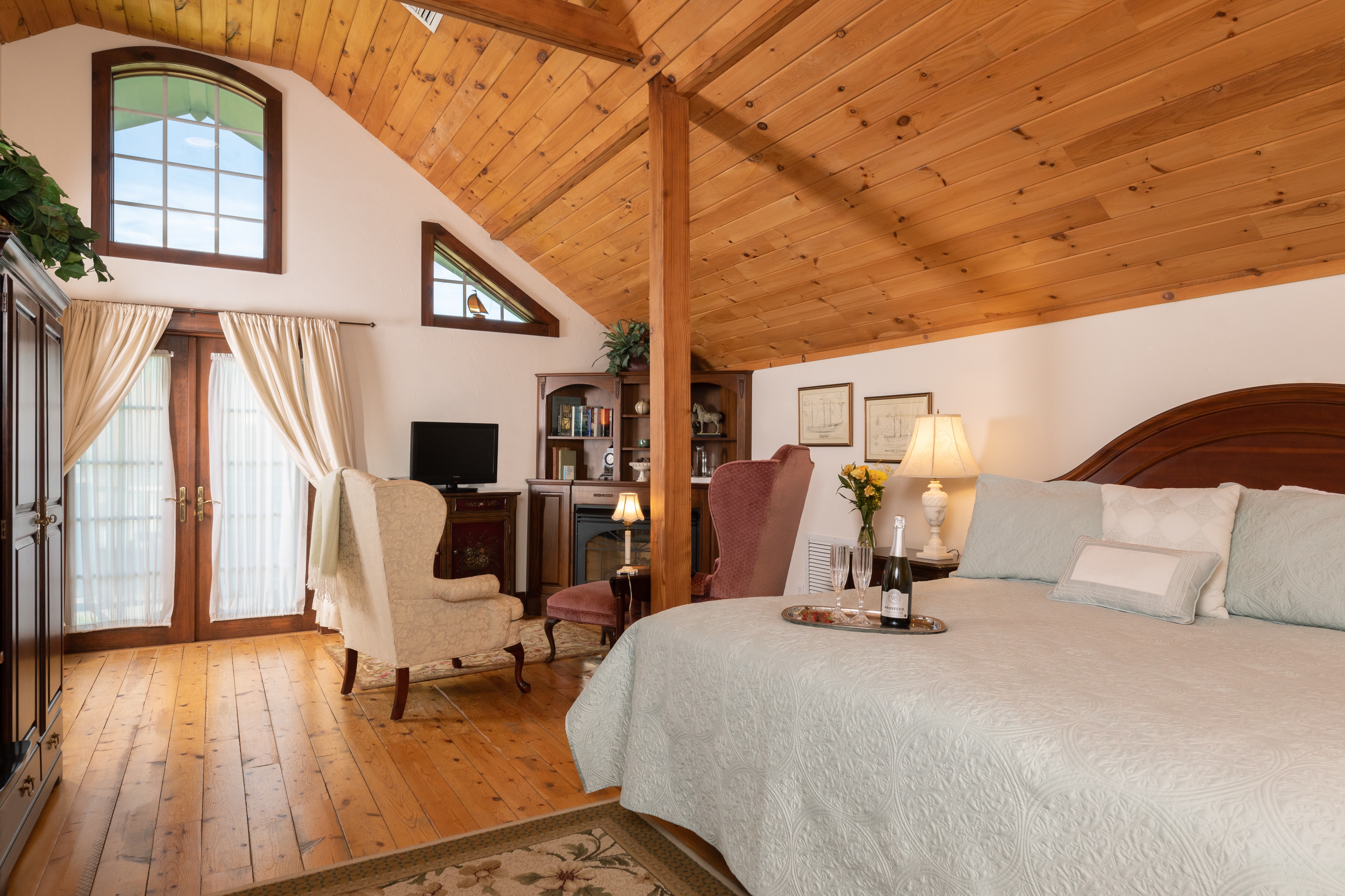 A spacious bedroom with a warm wood cathedral ceiling, hardwood floors, a king bed, and a cozy sitting area by the French doors.