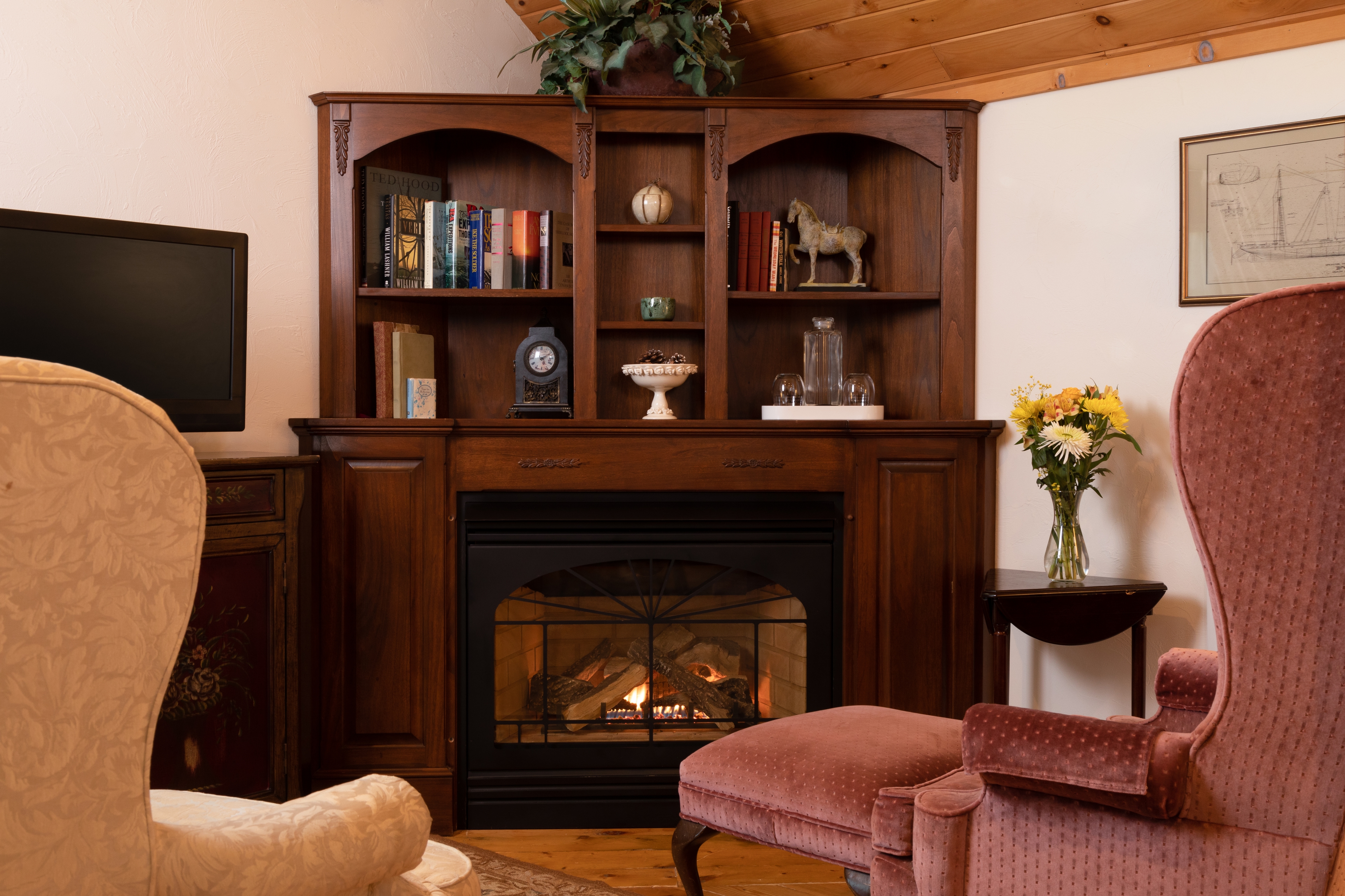 A cozy sitting area with a lit fireplace set in a dark wood bookcase mantel, flanked by two upholstered wingback chairs and an ottoman.