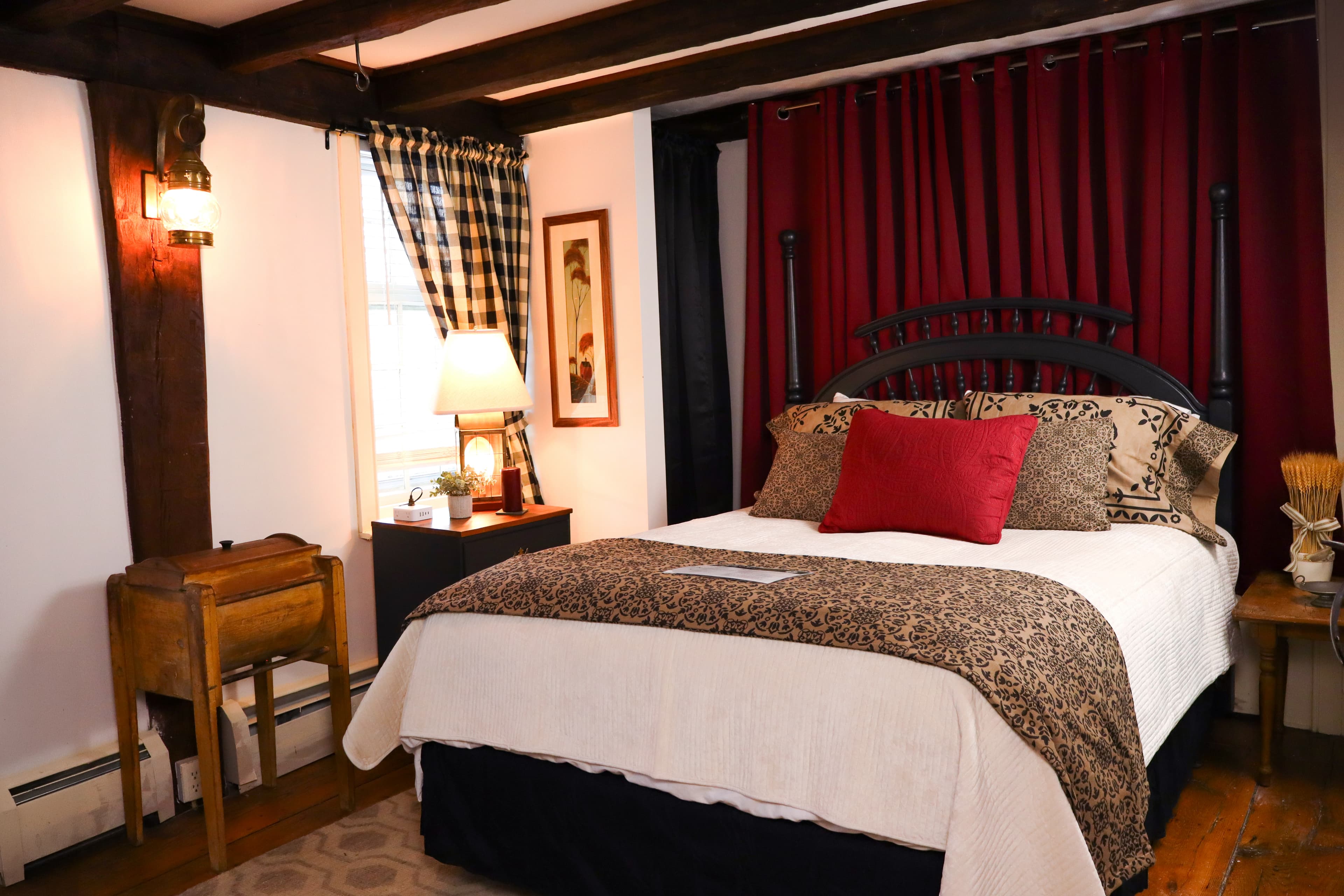 A warm cozy bedroom with dark wood exposed beams, a white quilt with a primitive style blanket, black nightstand and burgundy and plaid drapes