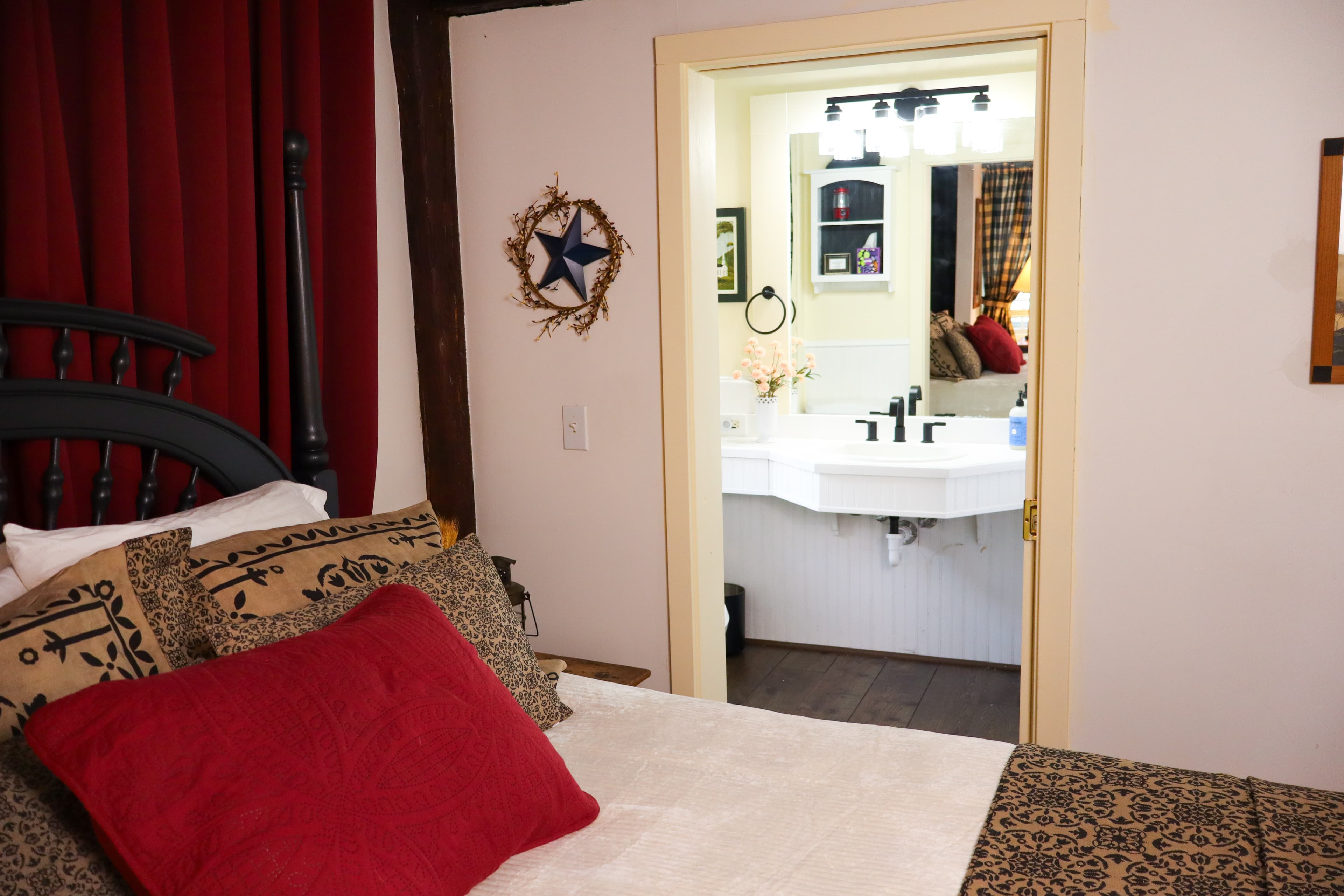 A warm cozy bedroom with dark wood exposed beams, a white quilt with a primitive style blanket and an open door leading to a brightly lit bathroom.