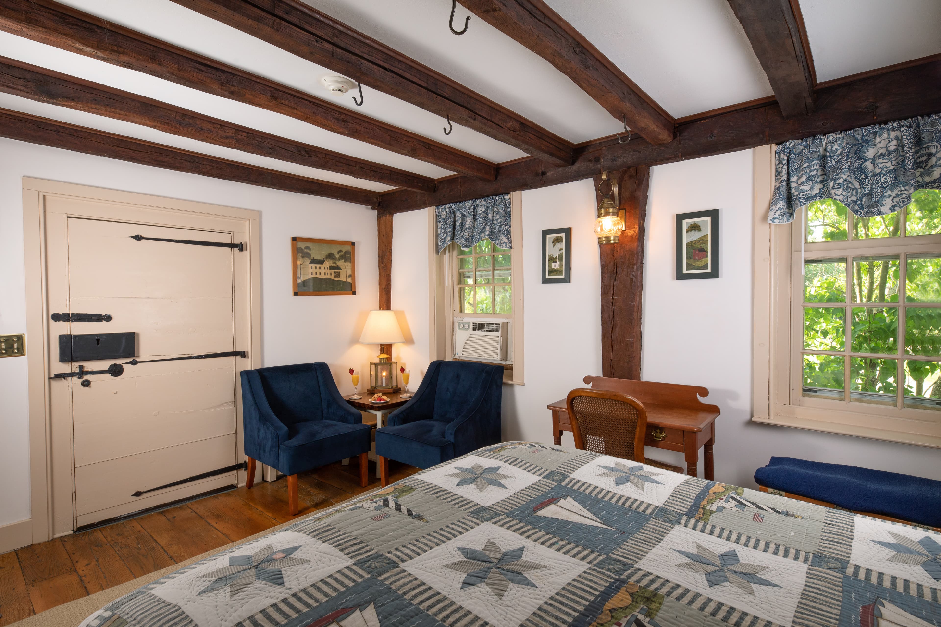 A quaint, nautical bedroom with a sailboat quilt, dark wood exposed beams, a Dutch door, and a small sitting area with blue velvet chairs.