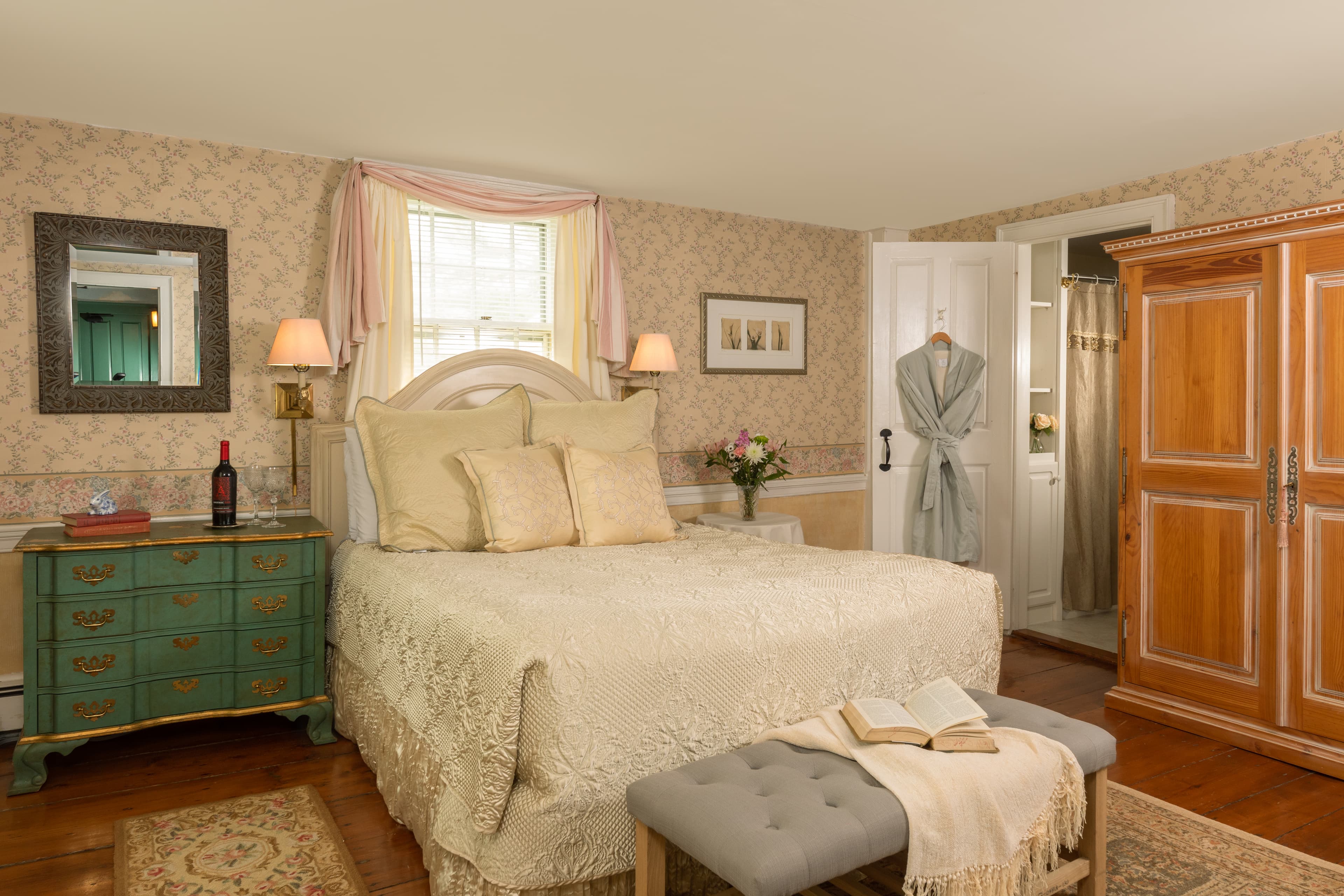A romantic bedroom featuring a large white bed, vintage patterned wallpaper, a teal dresser, and a light wood wardrobe with a robe hanging nearby.