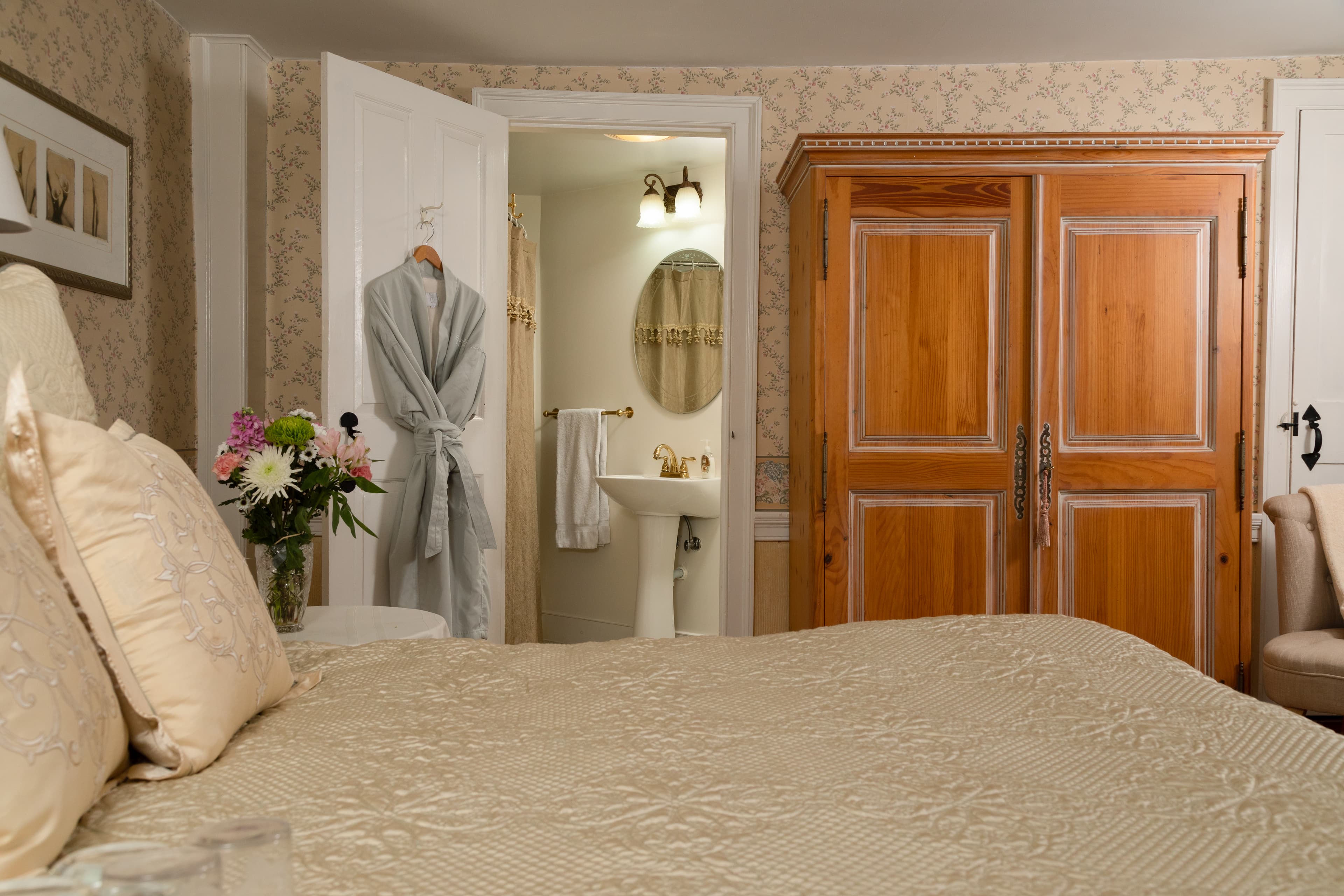 A bright, cozy bedroom with a large bed, light wood armoire, and an open doorway revealing a pedestal sink and mirror in the adjacent bathroom.