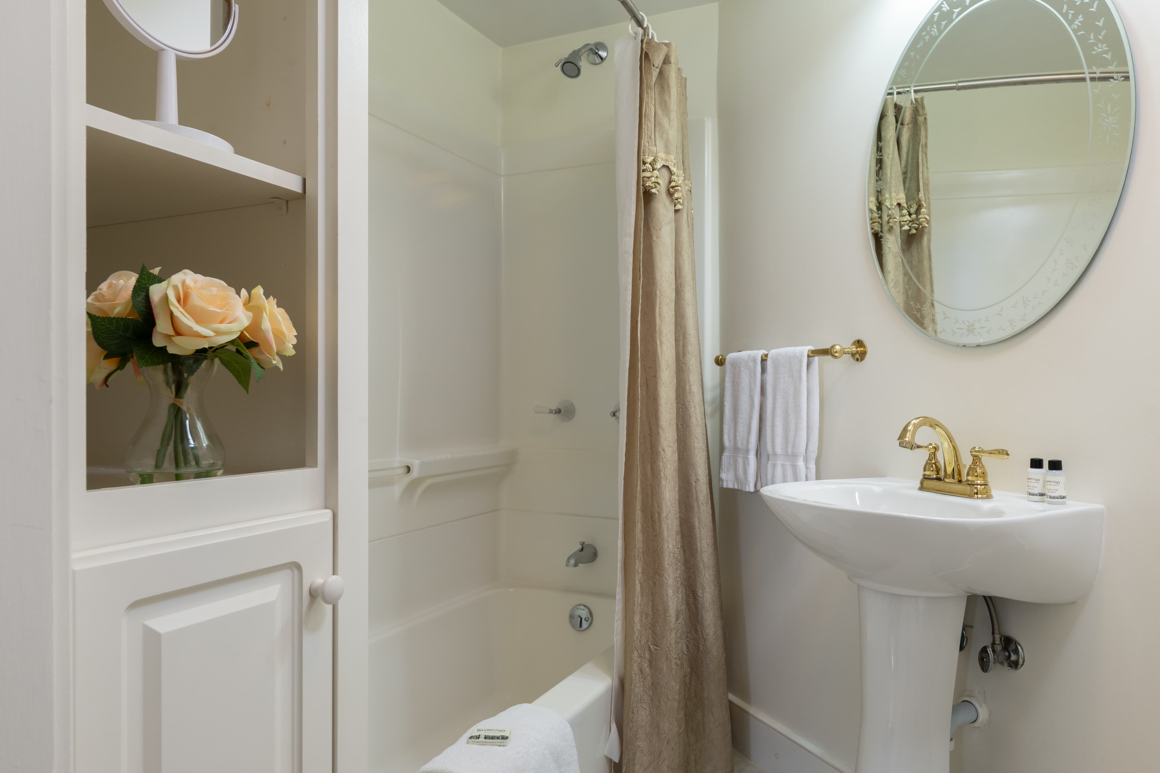 A small, charming bathroom featuring a pedestal sink with gold fixtures, a tub/shower combo, and built-in shelving with flowers.