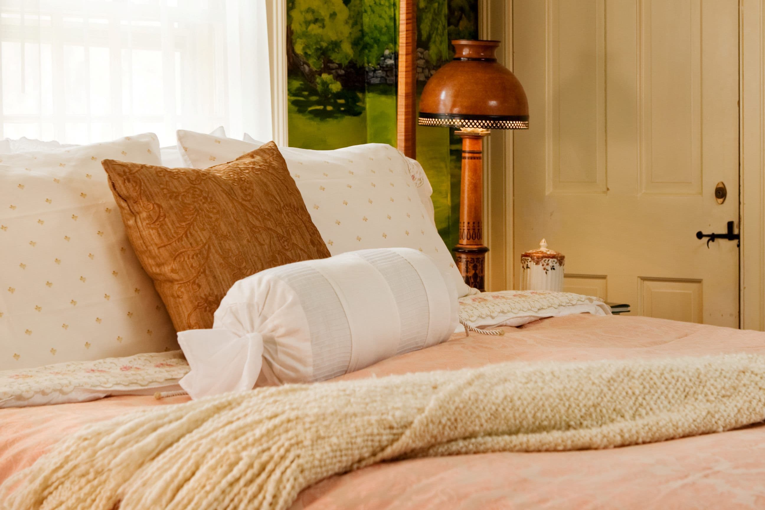 A close-up of a cozy bed with layered white and brown pillows, a white bolster, and a soft pink/peach blanket.