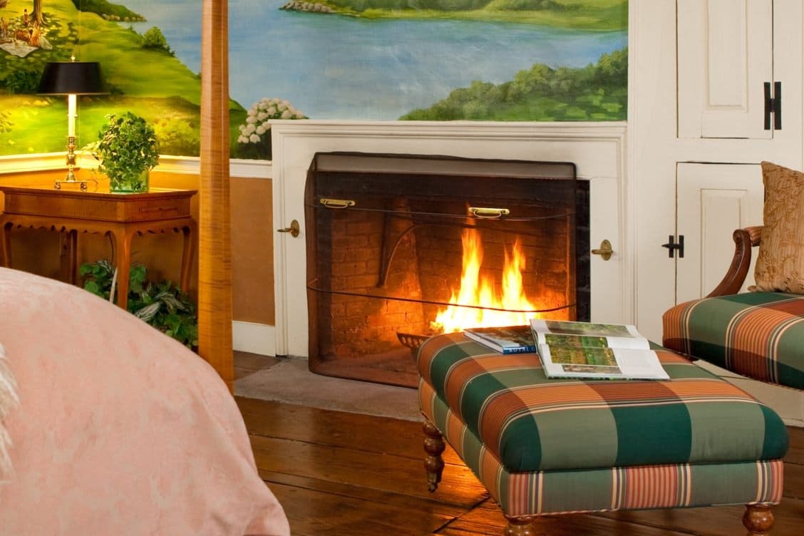 A romantic bedroom scene with a lit fireplace, a plaid ottoman, and a hand-painted landscape mural, creating a cozy and unique ambiance.