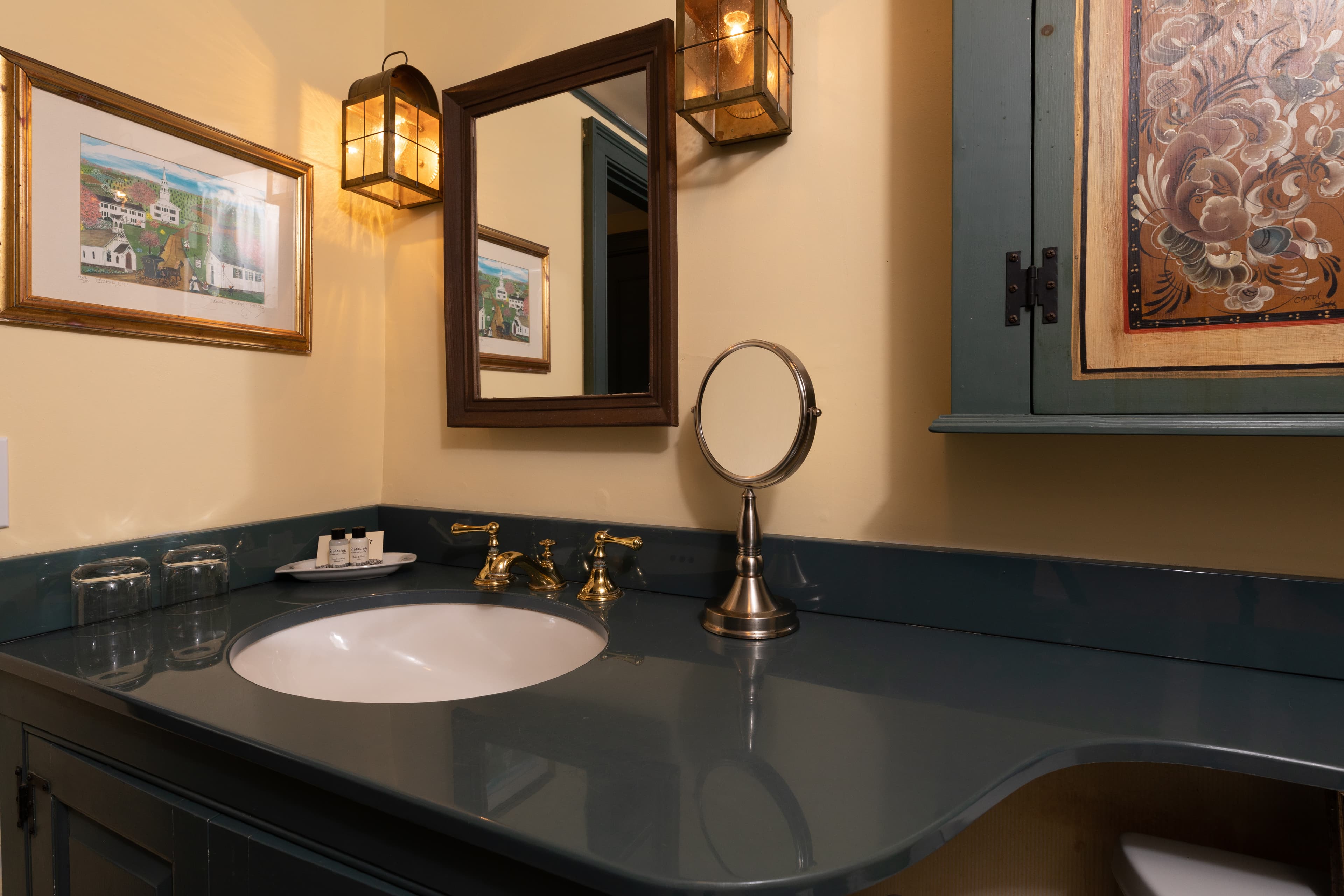 A classic bathroom vanity featuring a dark teal countertop and cabinet, a wood-framed mirror with lantern sconces, and brass fixtures.