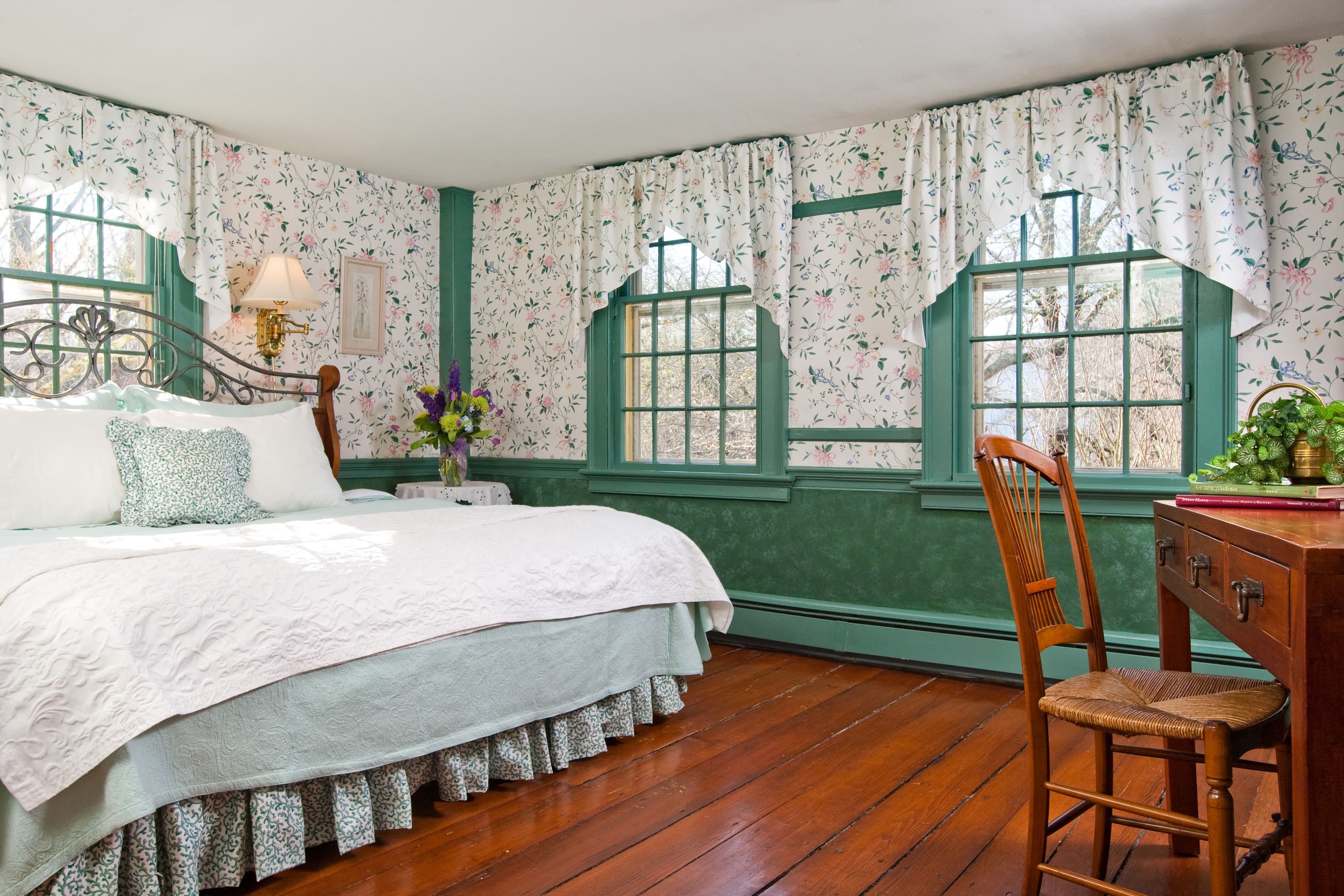 A quaint, charming bedroom with floral wallpaper, bright green wainscoting and window trim, a large bed, and hardwood floors.