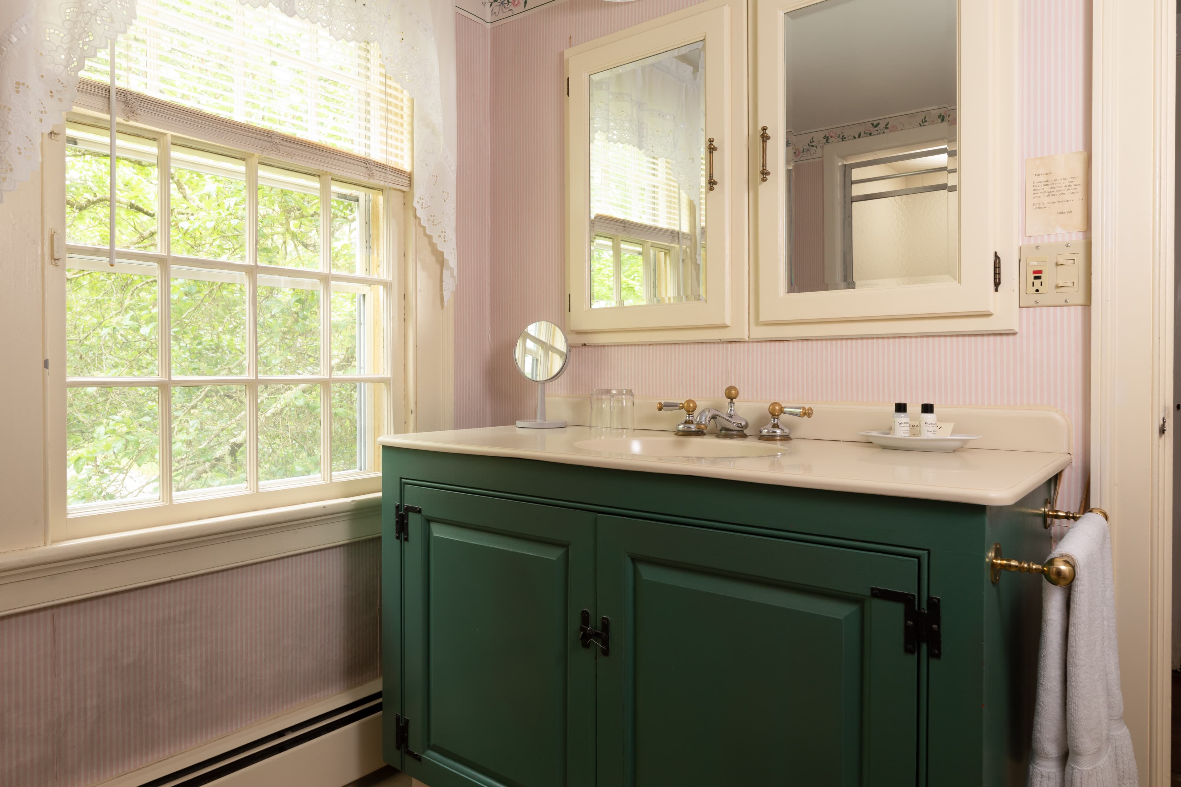 A bright, quaint bathroom featuring a bold green vanity with a white countertop, a mirrored cabinet, and a large window with sheer curtains.