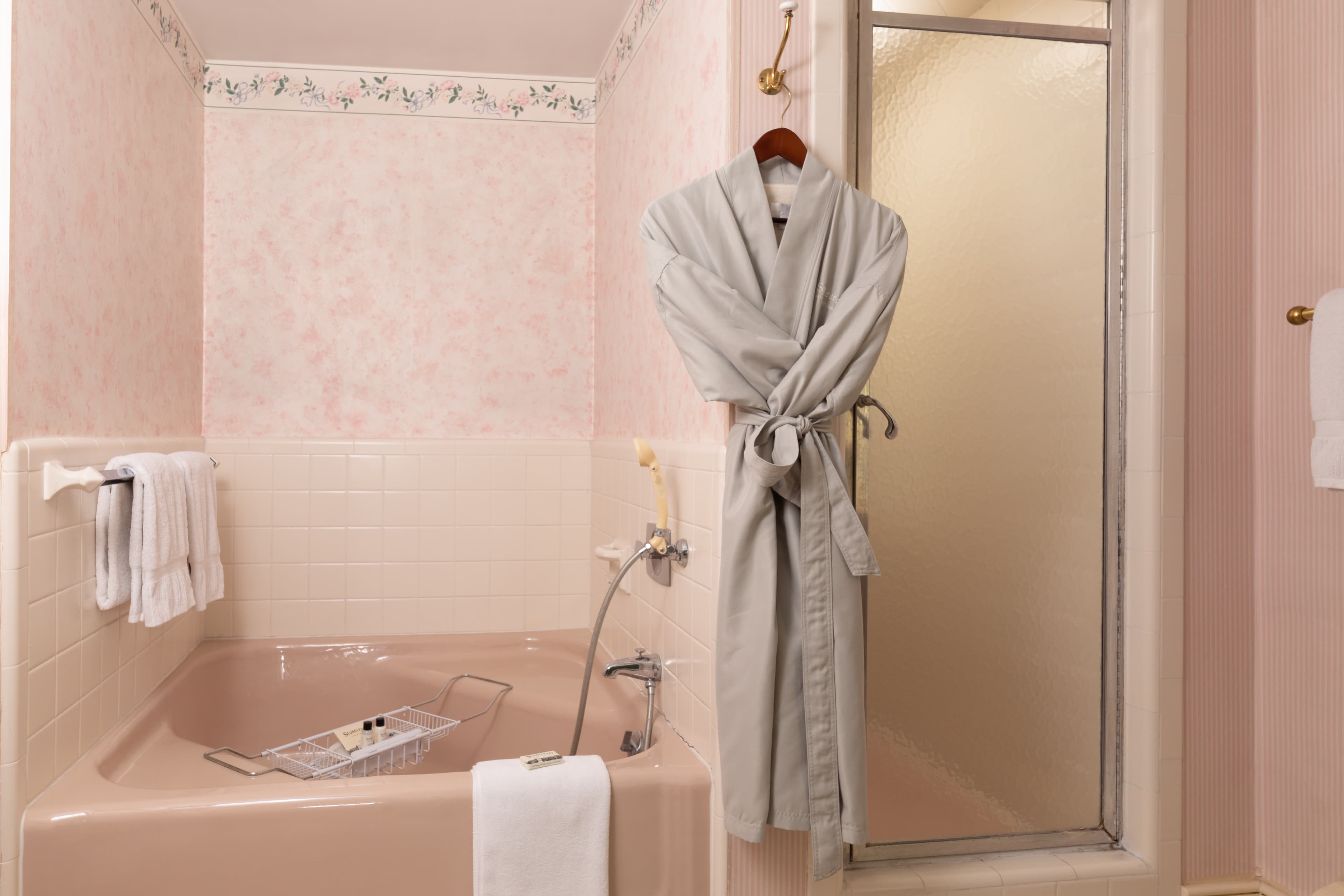 A vintage-style pink bathroom featuring a built-in tub, a glass shower stall, and a cozy gray bathrobe hanging on the wall.