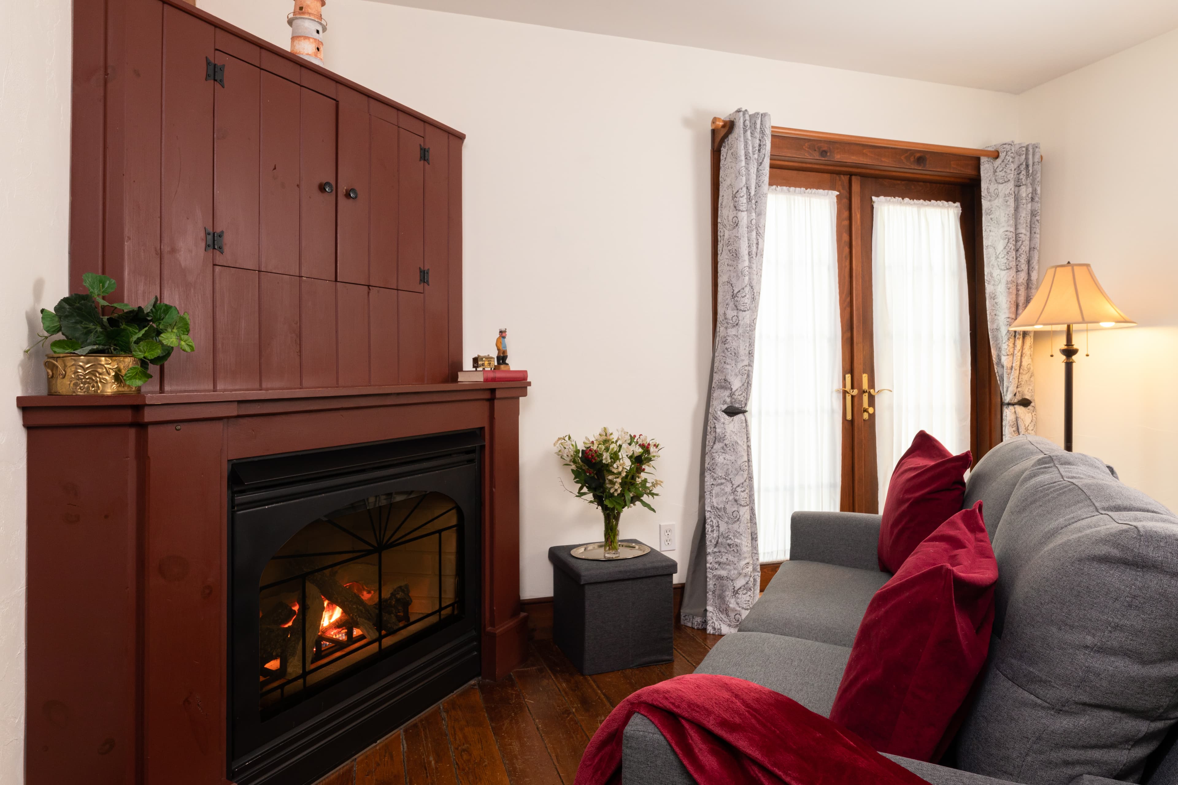 A cozy sitting area with a lit fireplace set in a dark red cabinet, a gray sofa with crimson pillows, and French doors to the right.