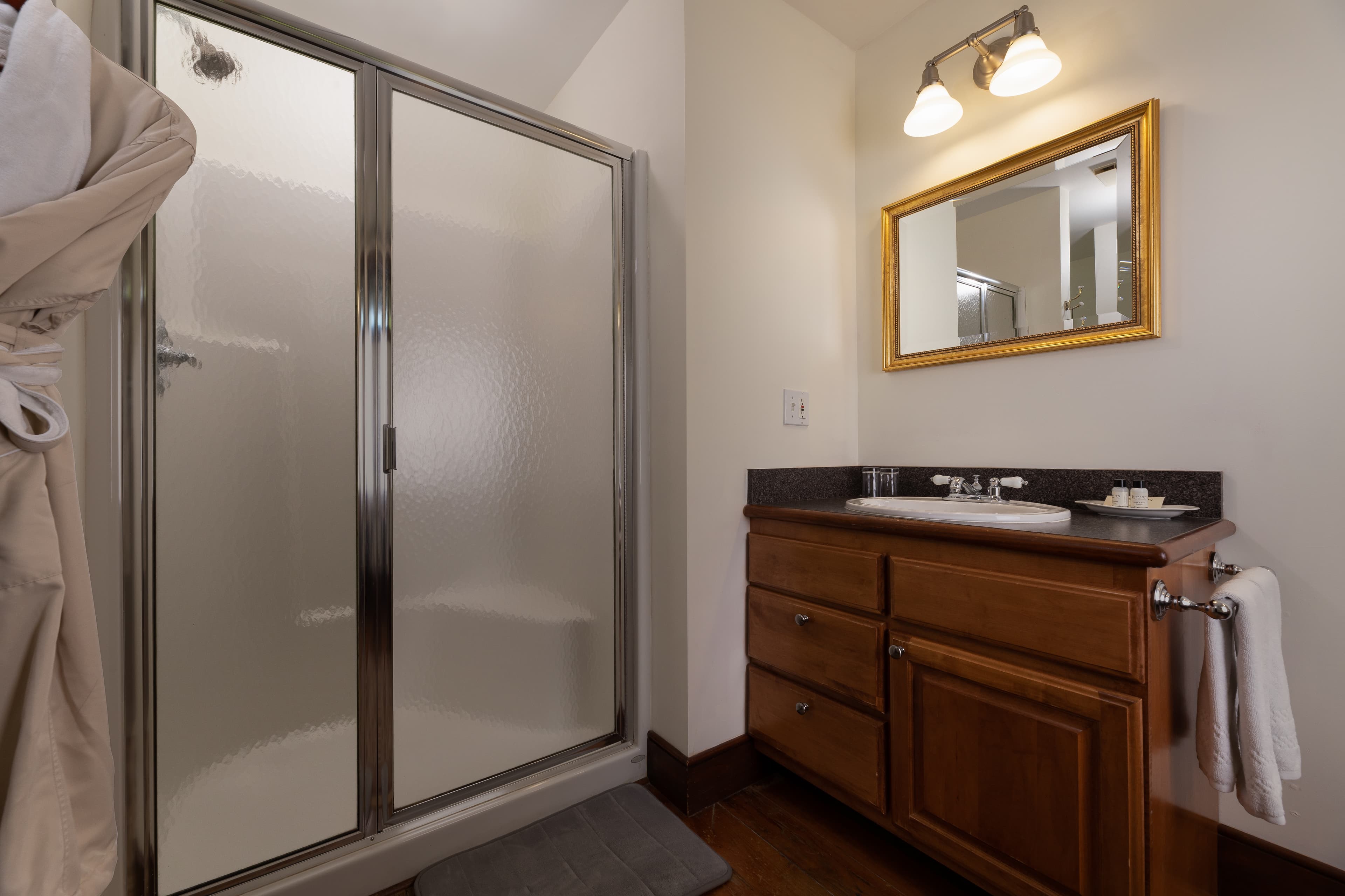 A bright bathroom featuring a glass-enclosed walk-in shower with a robe hanging nearby, and a warm wood vanity with a gold-framed mirror.