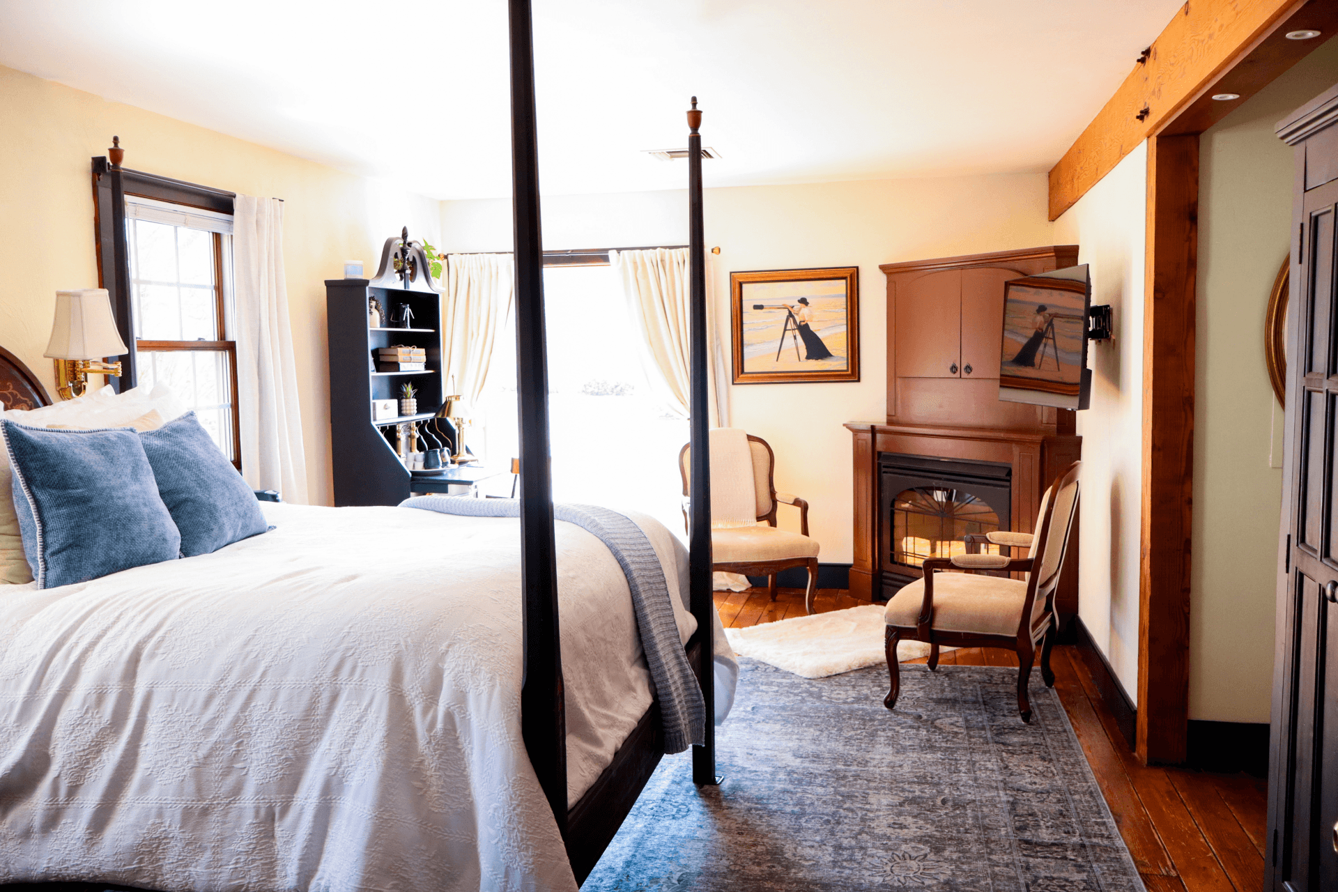 A cozy, elegant bedroom with a dark wood four-poster bed, a fireplace, comfortable armchairs, and floral curtains.