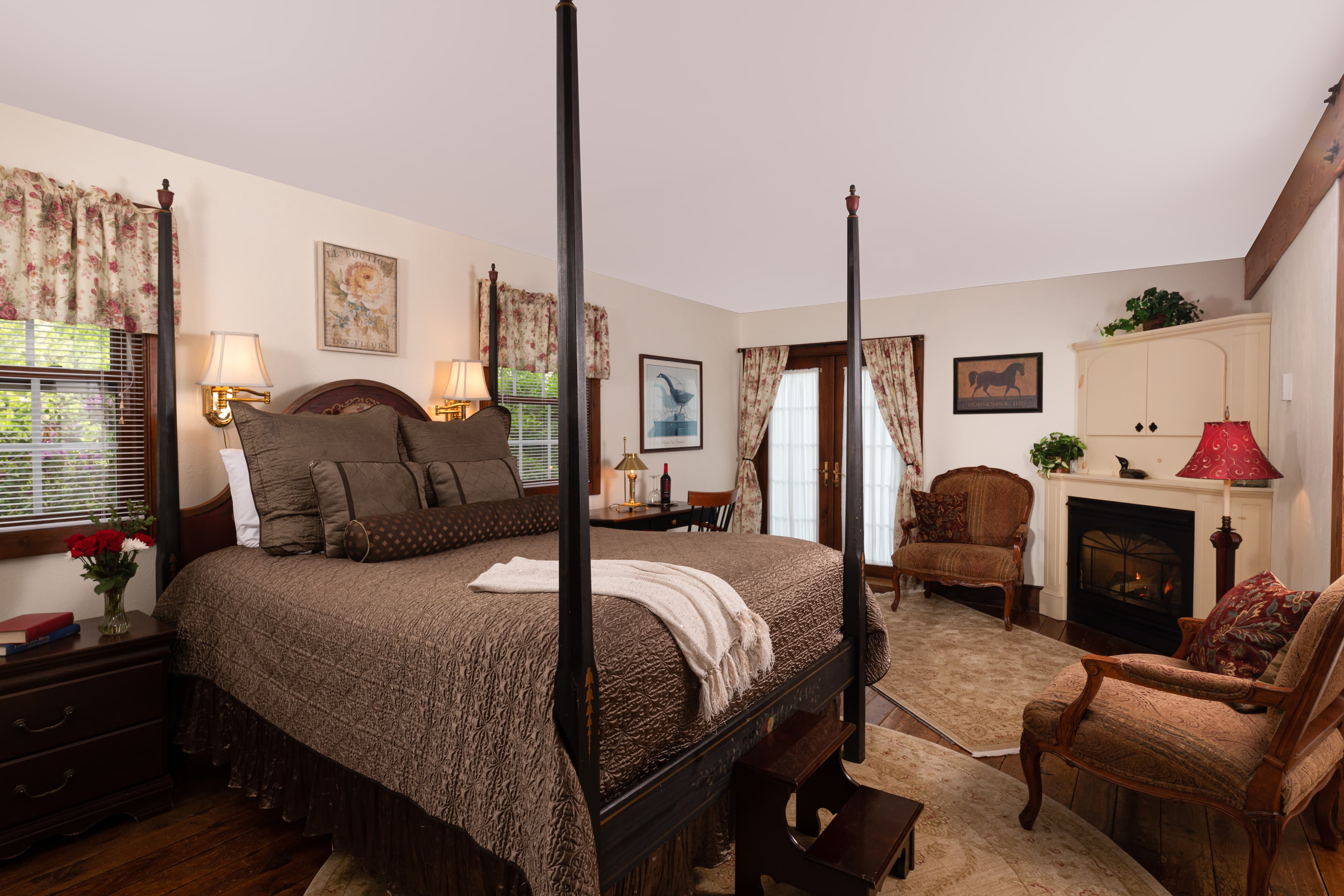A cozy, elegant bedroom with a dark wood four-poster bed, a fireplace, comfortable armchairs, and floral curtains.