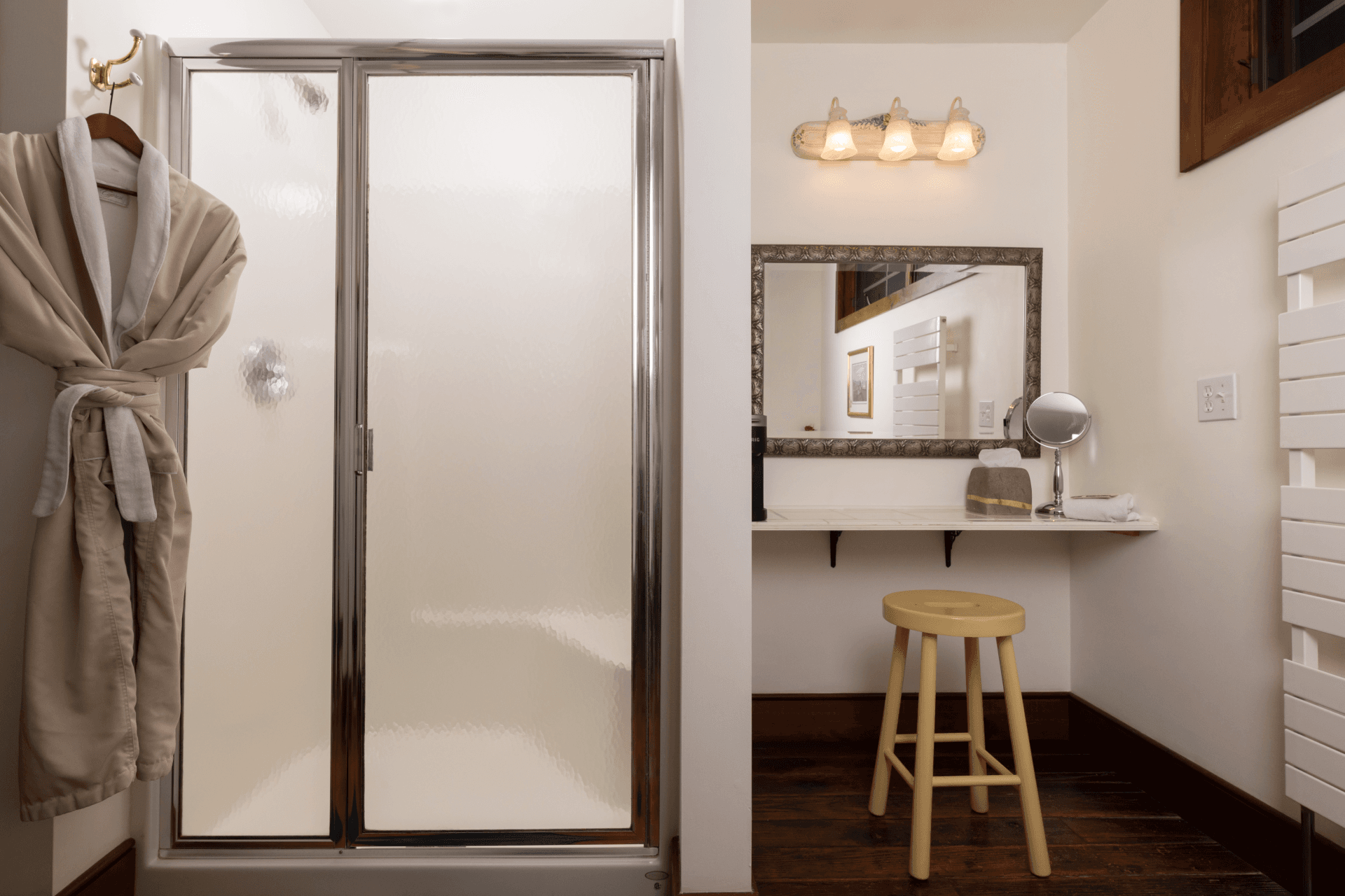 Glass walk-in shower and vanity area with mirror, stool, and plush robe in the Briar Rose room bathroom at Stonecroft Country Inn, featuring warm lighting and elegant country inn décor.