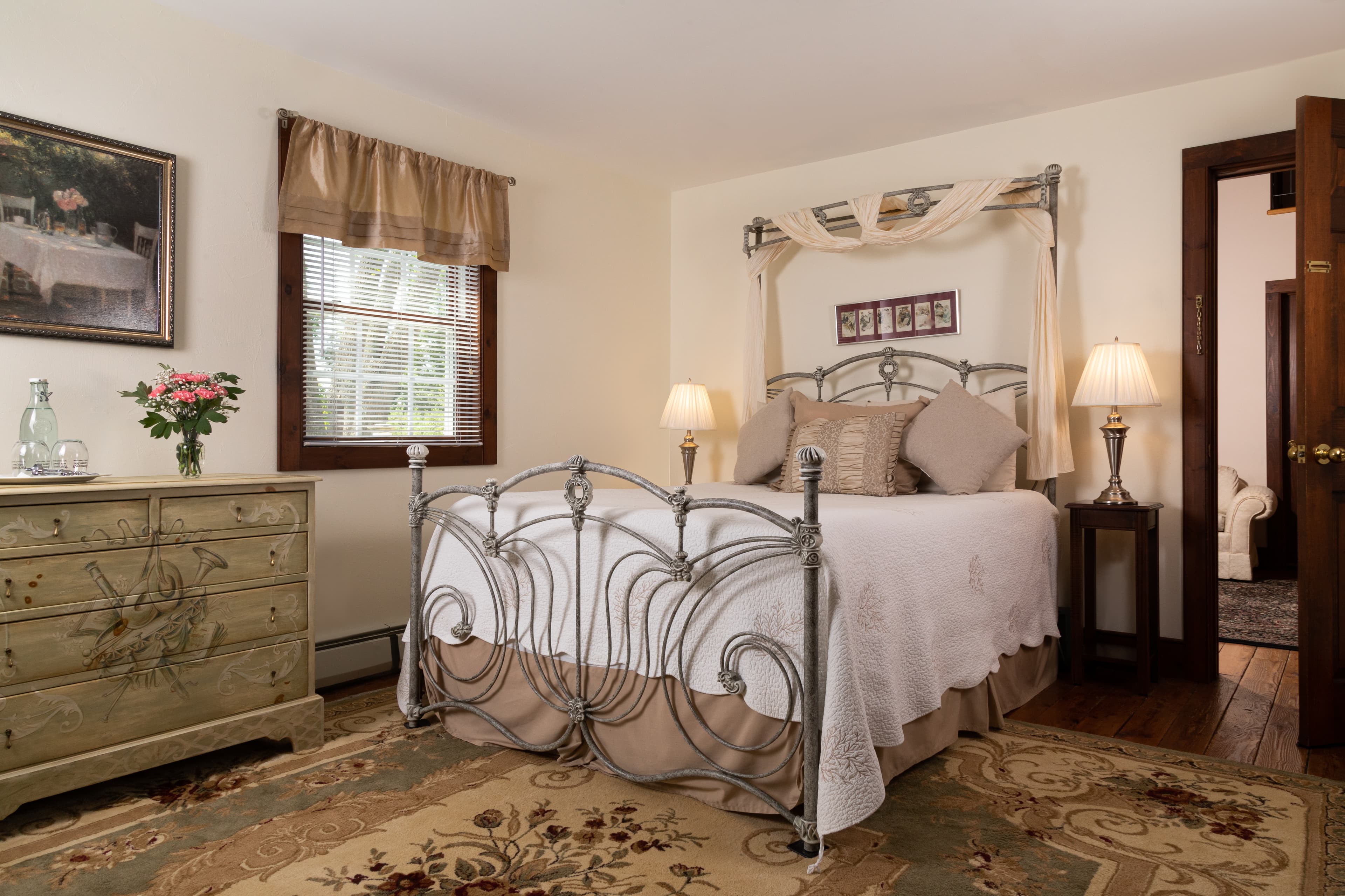 A bright, romantic bedroom with a scrollwork metal bed frame, a light green antique dresser, and a doorway leading to an adjoining room.