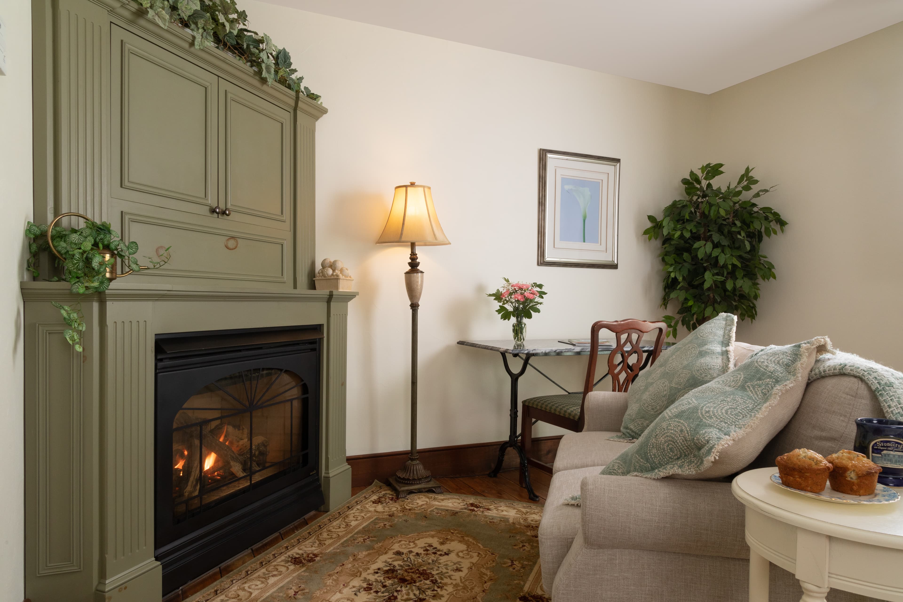 A cozy sitting area featuring a lit fireplace set in a sage-green mantel, a comfortable sofa with pillows, and a small table with flowers.