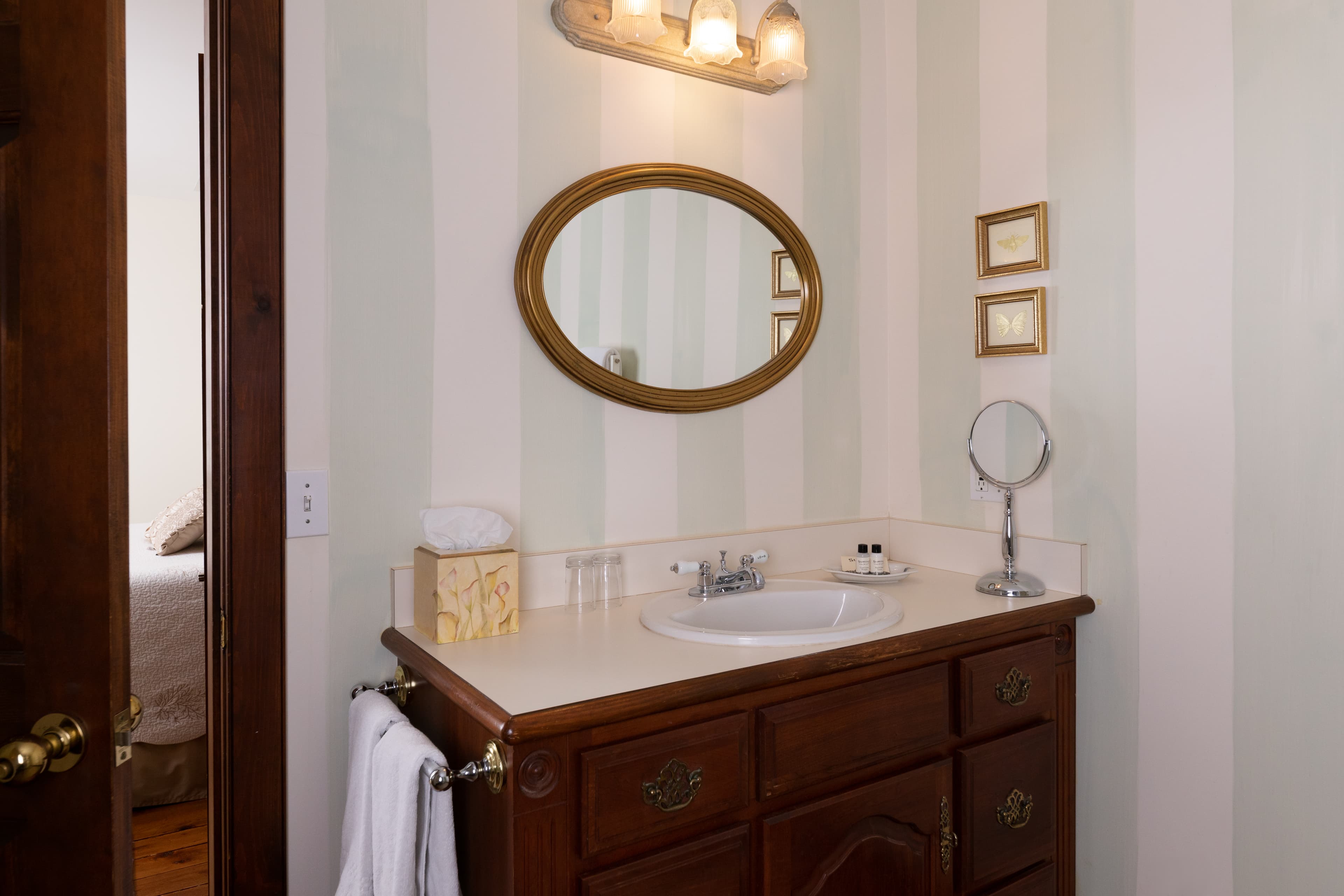 A classic bathroom vanity featuring a dark wood cabinet, a countertop sink, a gold-framed oval mirror, and striped wallpaper.