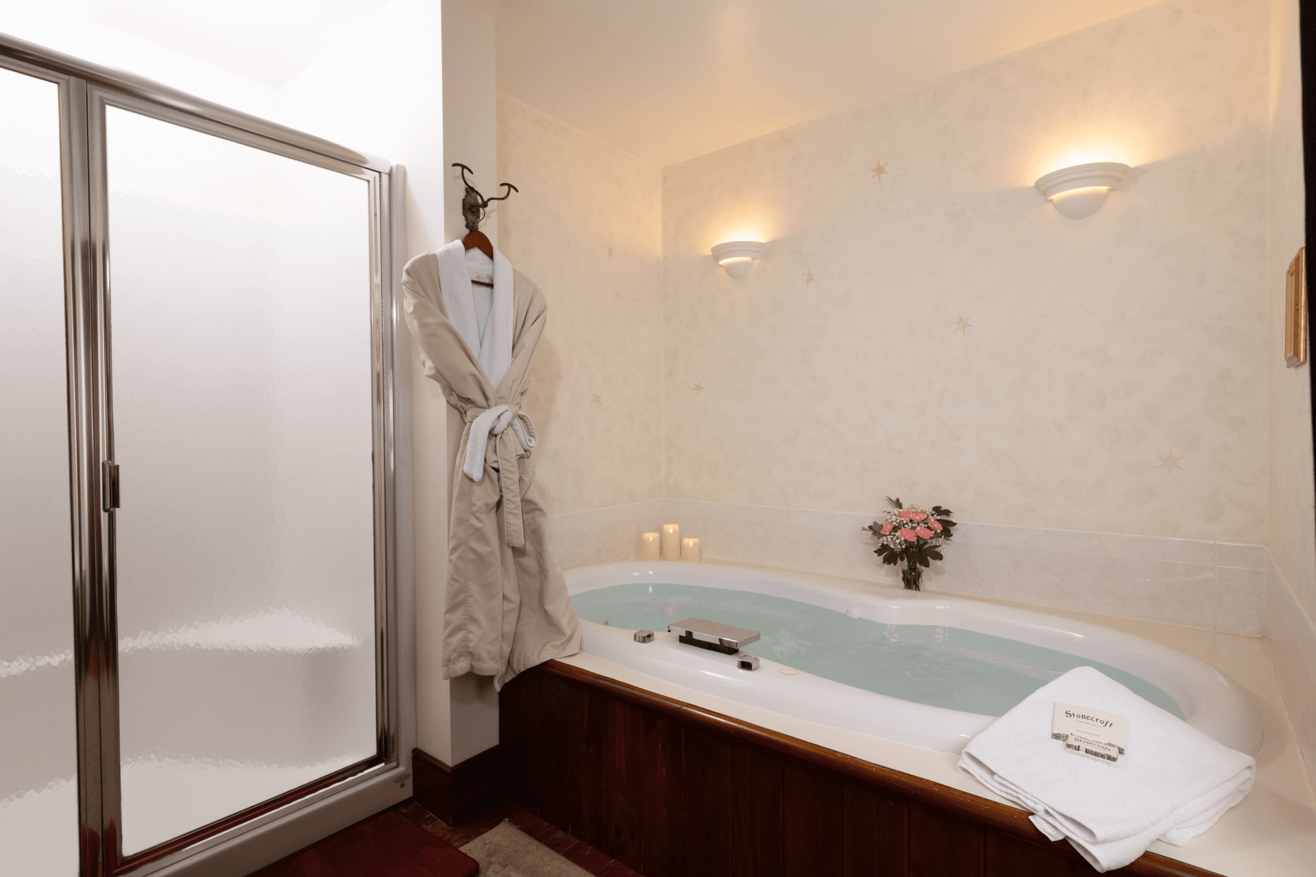 Luxury jetted soaking tub in the Lily Suite at Stonecroft Inn, a boutique bed and breakfast in Ledyard, CT, featuring a whirlpool tub with wood surround, lit candles, fresh pink roses, plush white robes, and a separate glass-door shower.