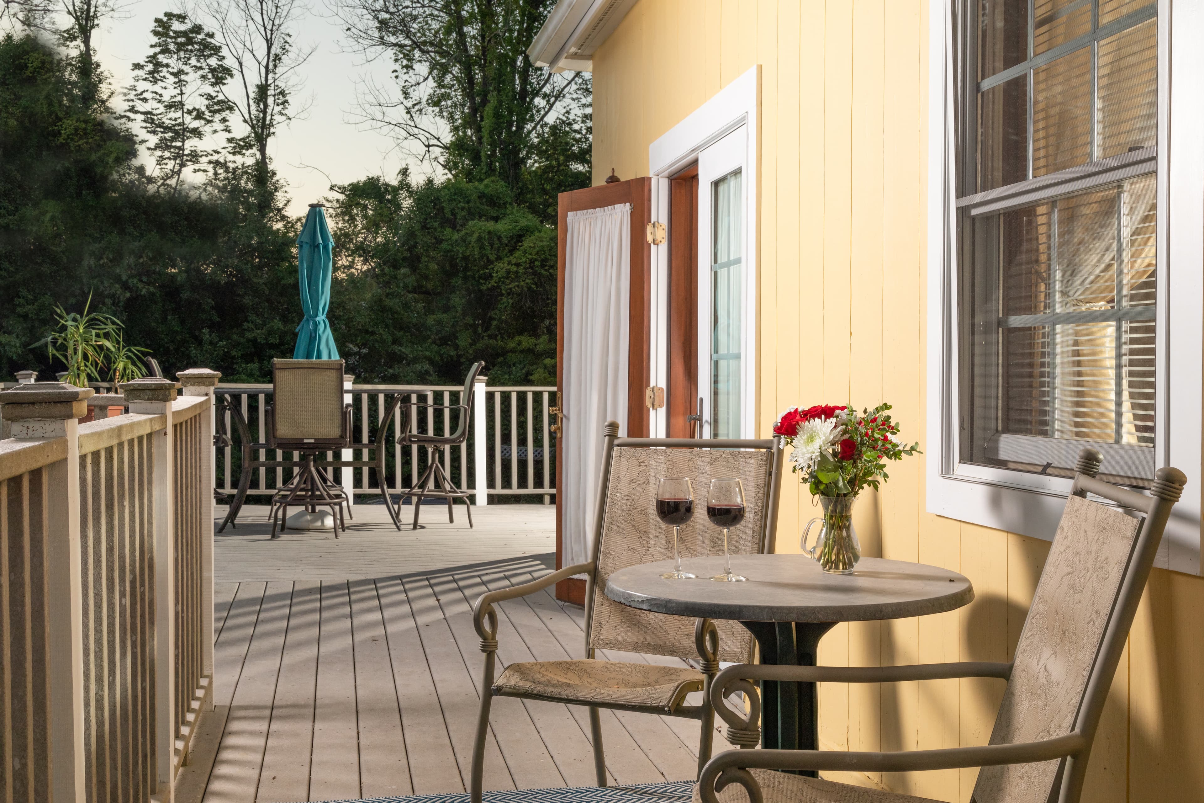 A charming outdoor deck with yellow siding, a small bistro table for two with wine glasses, and a view of surrounding greenery.
