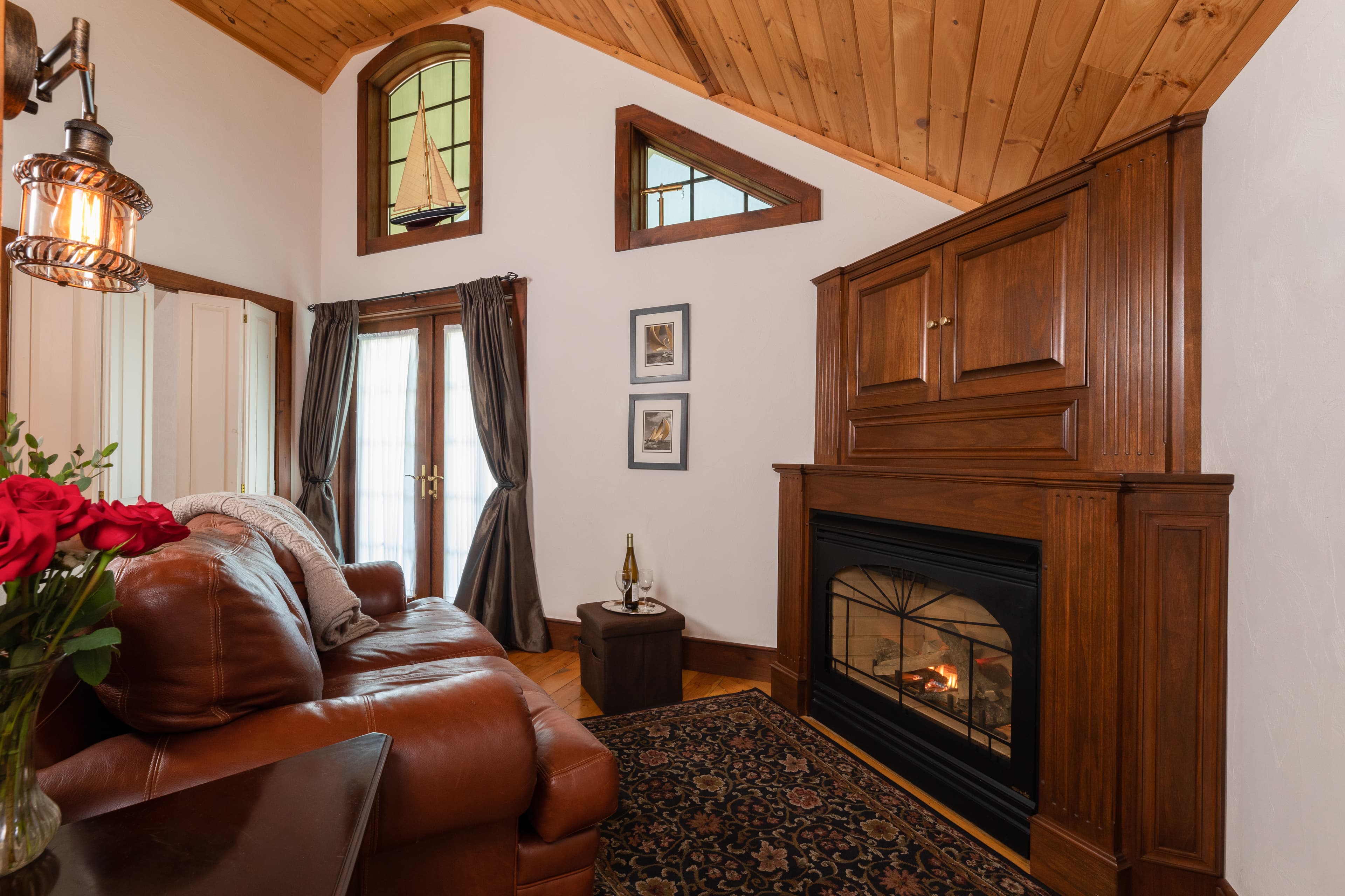 A cozy loft sitting area with a warm wood cathedral ceiling, a lit fireplace in a wood mantel, a brown leather sofa, and French doors.