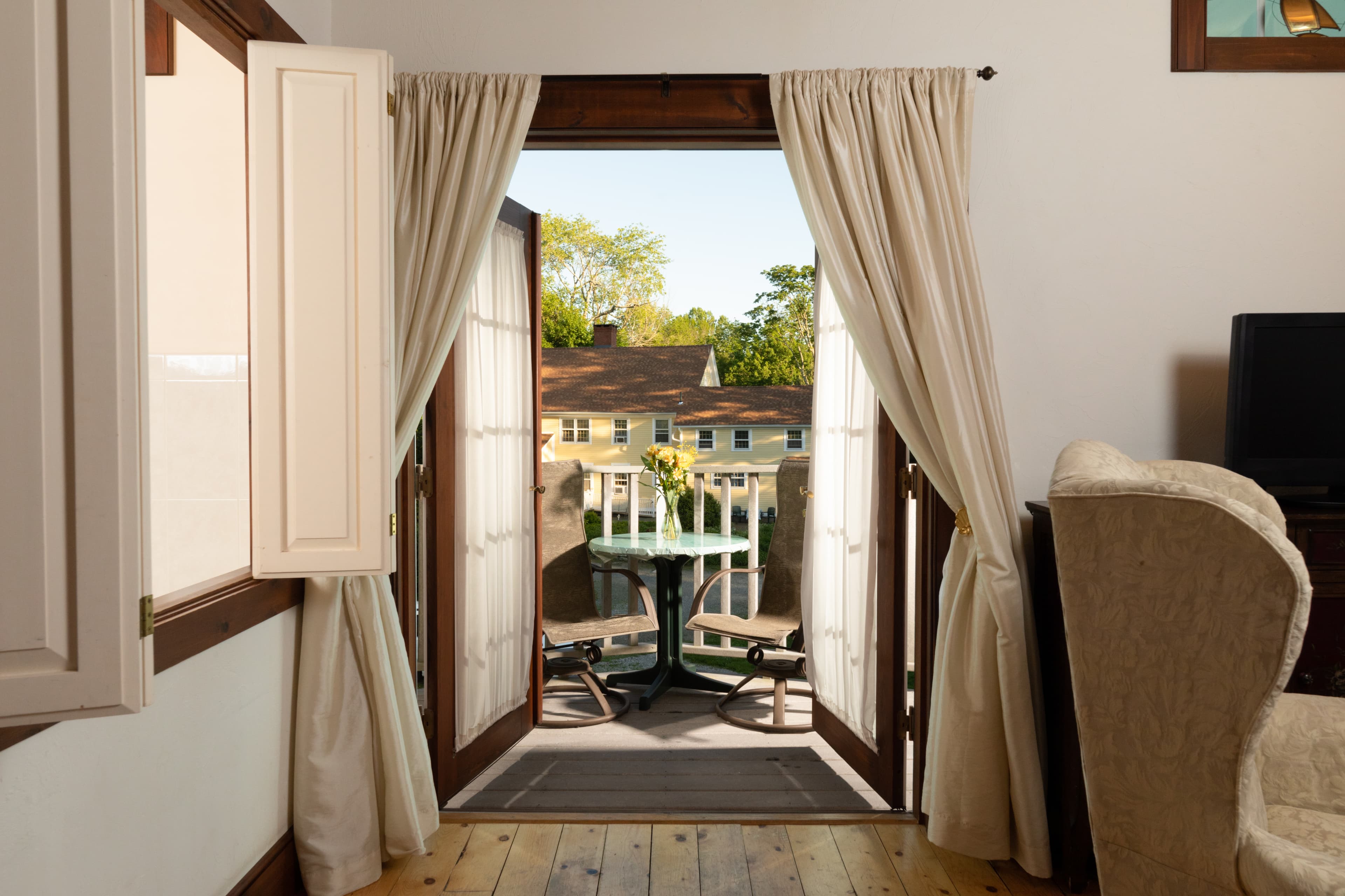Bright French doors open to a private balcony with a small bistro table, offering a sunny view of a charming yellow building and trees.