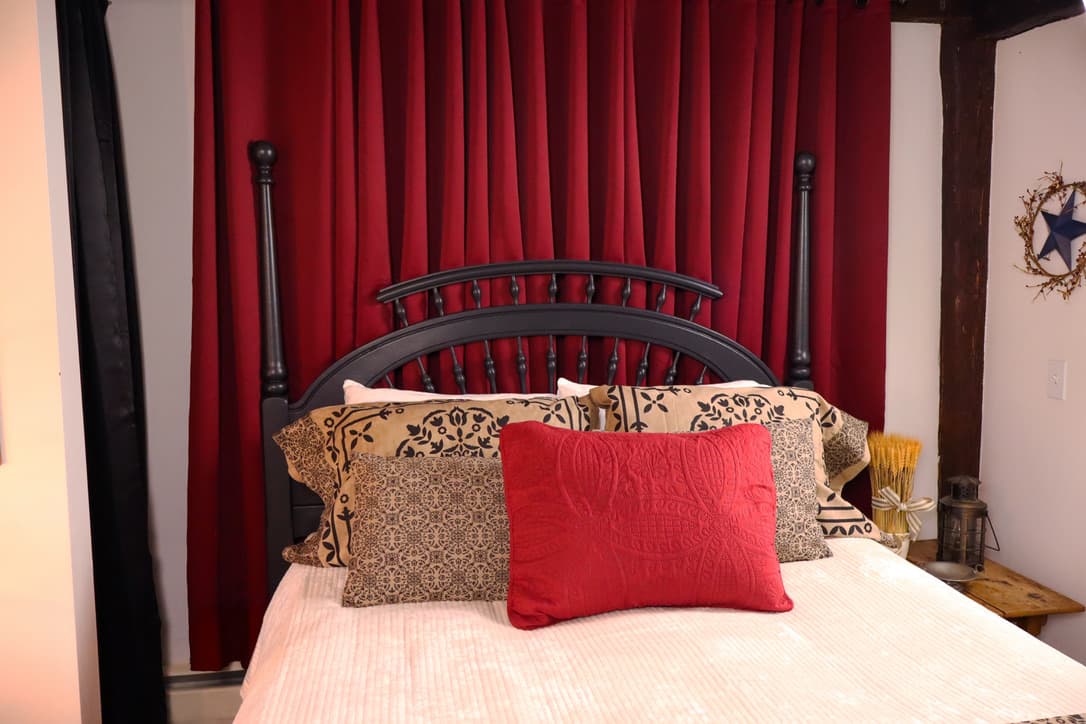 A black bed frame with a white quilt and burgundy throw pillow and burgundy drapes