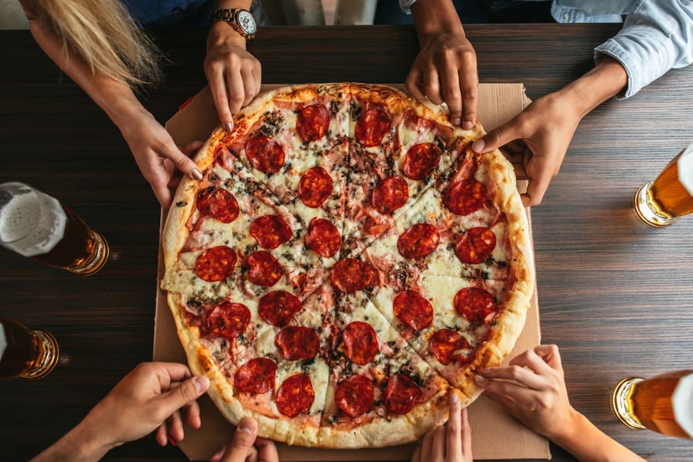 A group of hands reaching for a large pepperoni pizza on a table with beers nearby.